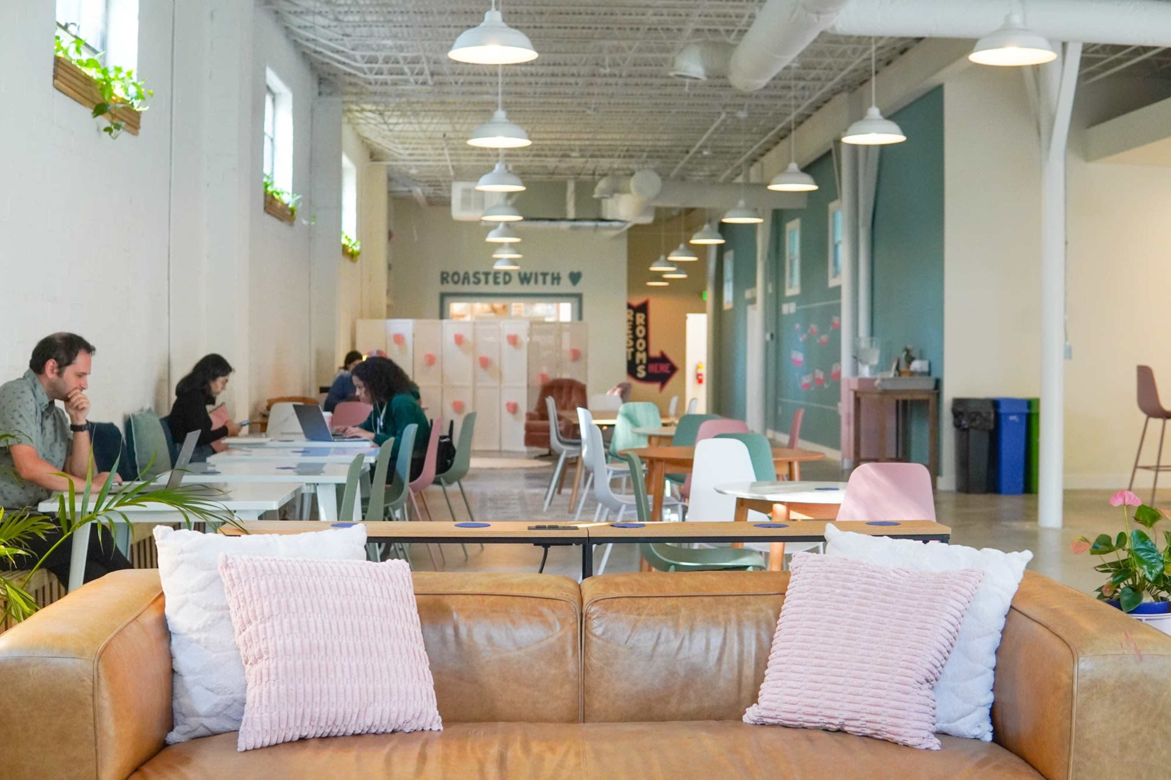 A cozy café interior features a brown leather sofa with pink cushions in the foreground, while patrons sit at various tables and work on laptops in a bright, open space.