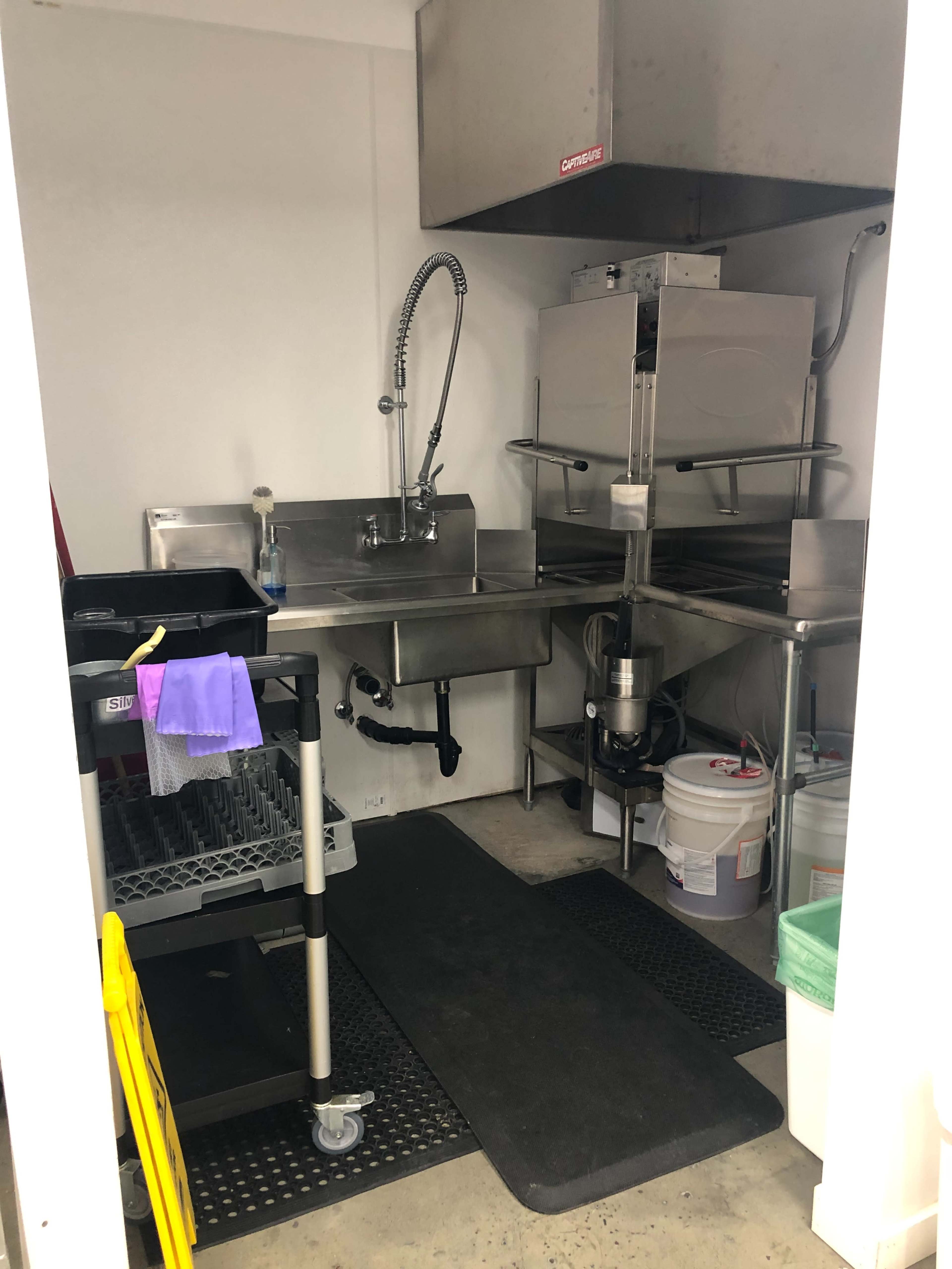A small kitchen area equipped with a stainless steel sink, a dish rack, cleaning supplies, and a floor mat.