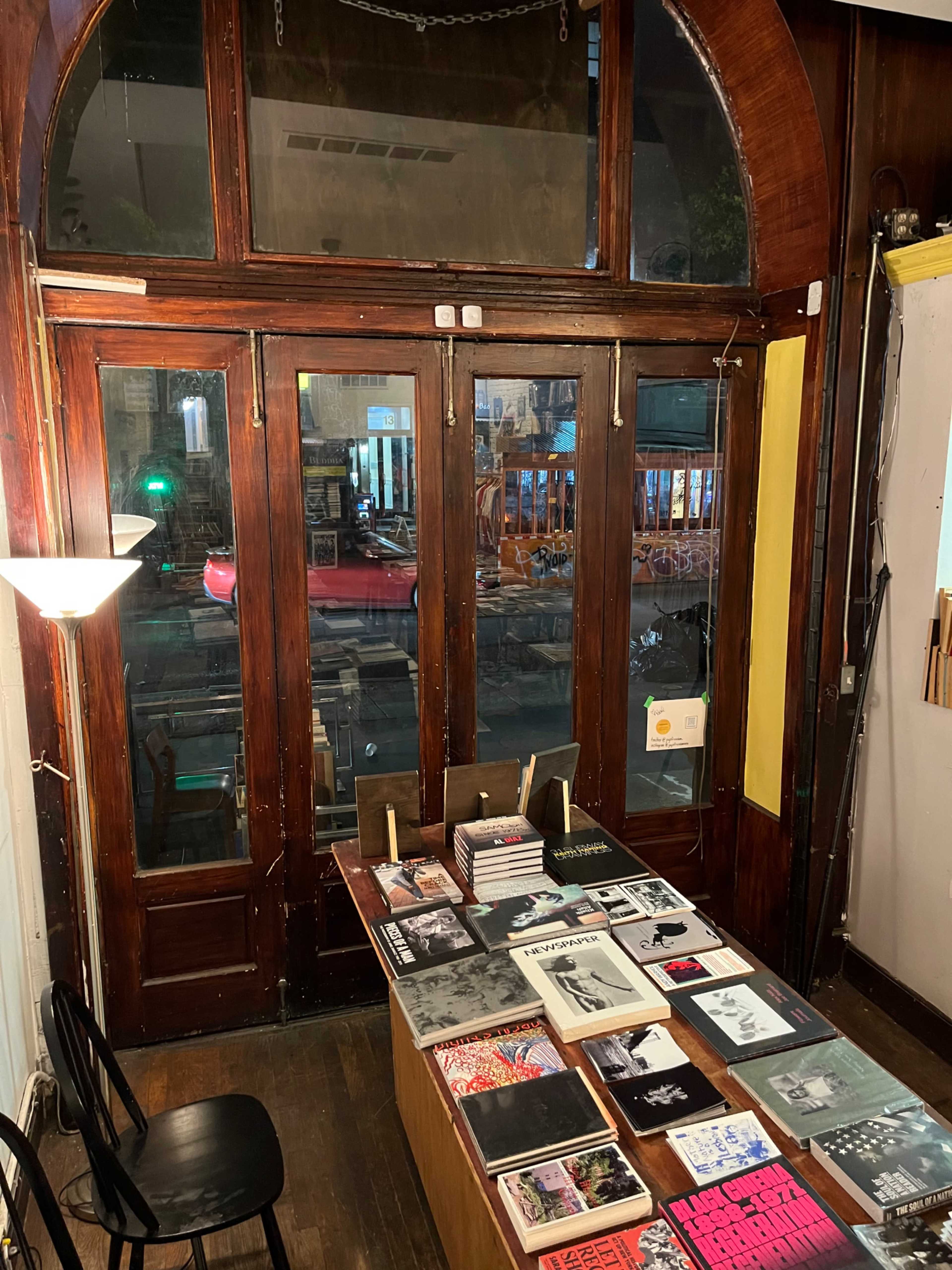 A cozy interior of a bookshop with a wooden table displaying various books and magazines, and large windows exposing a night-time street view.