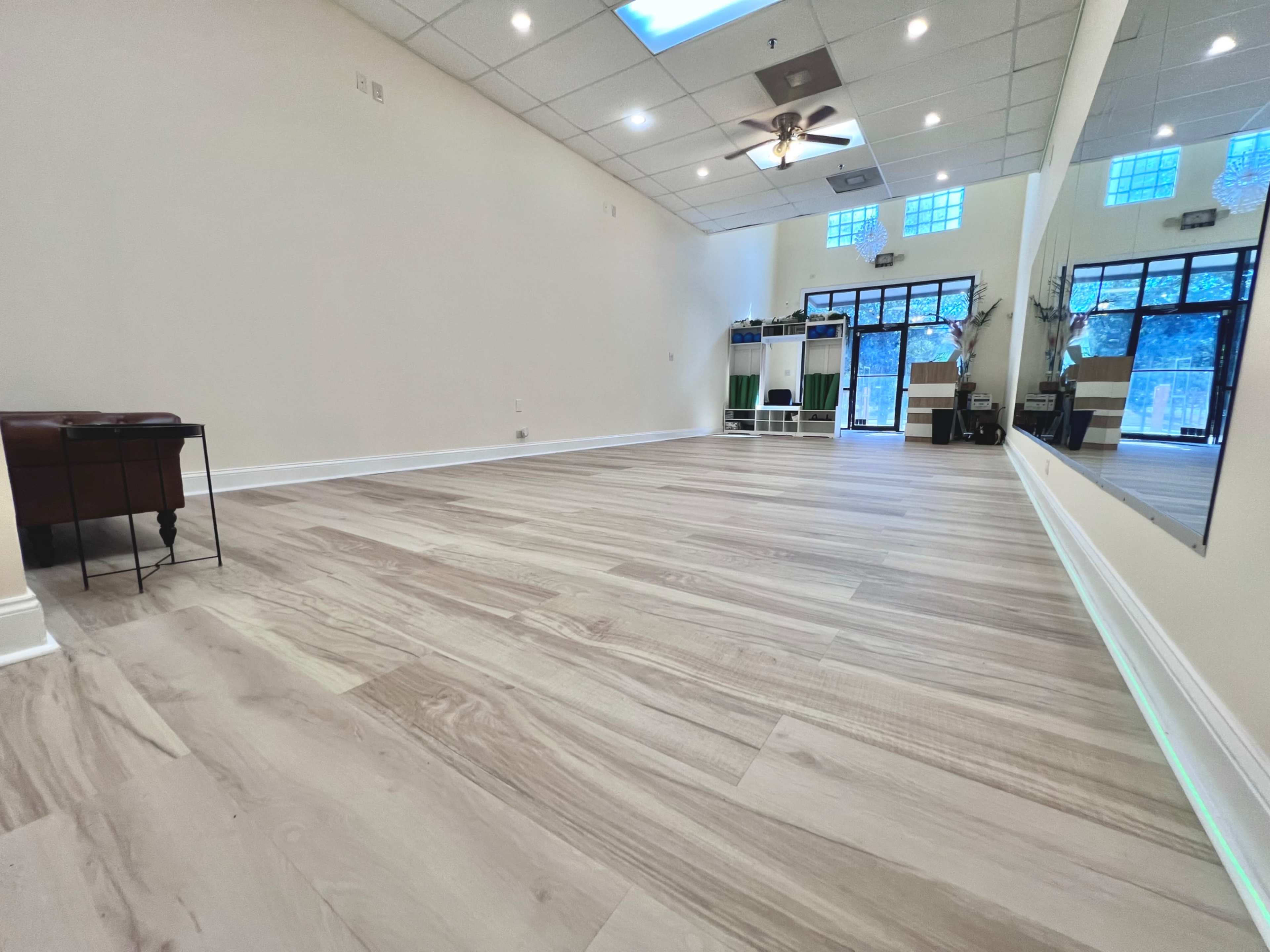 The image shows a spacious, well-lit indoor room with light wood flooring and large windows.