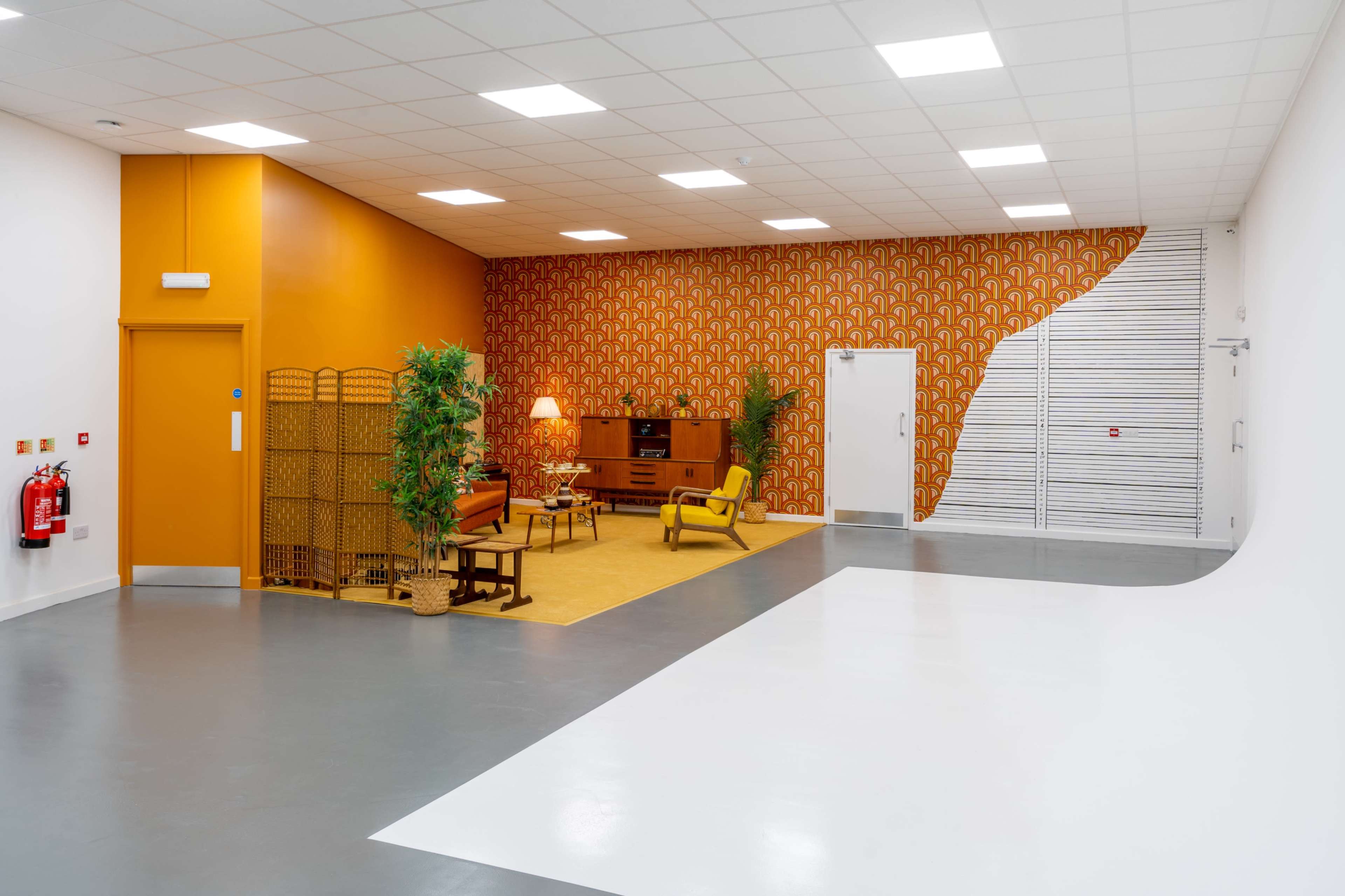 The image shows a spacious room with a retro-themed sitting area featuring an orange accent wall, patterned wallpaper, and yellow furniture.