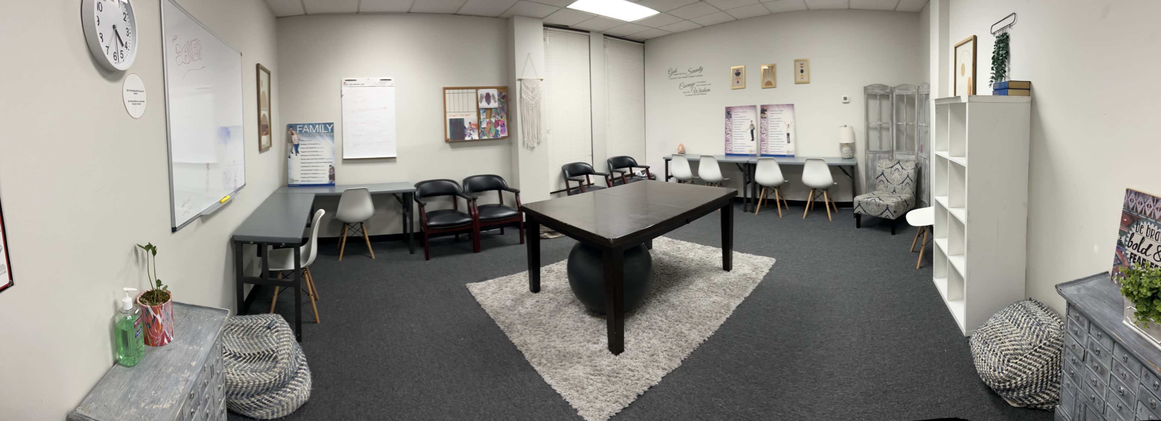 The image shows a spacious meeting room featuring a large black table surrounded by various chairs, a whiteboard, and shelves with decorative items.