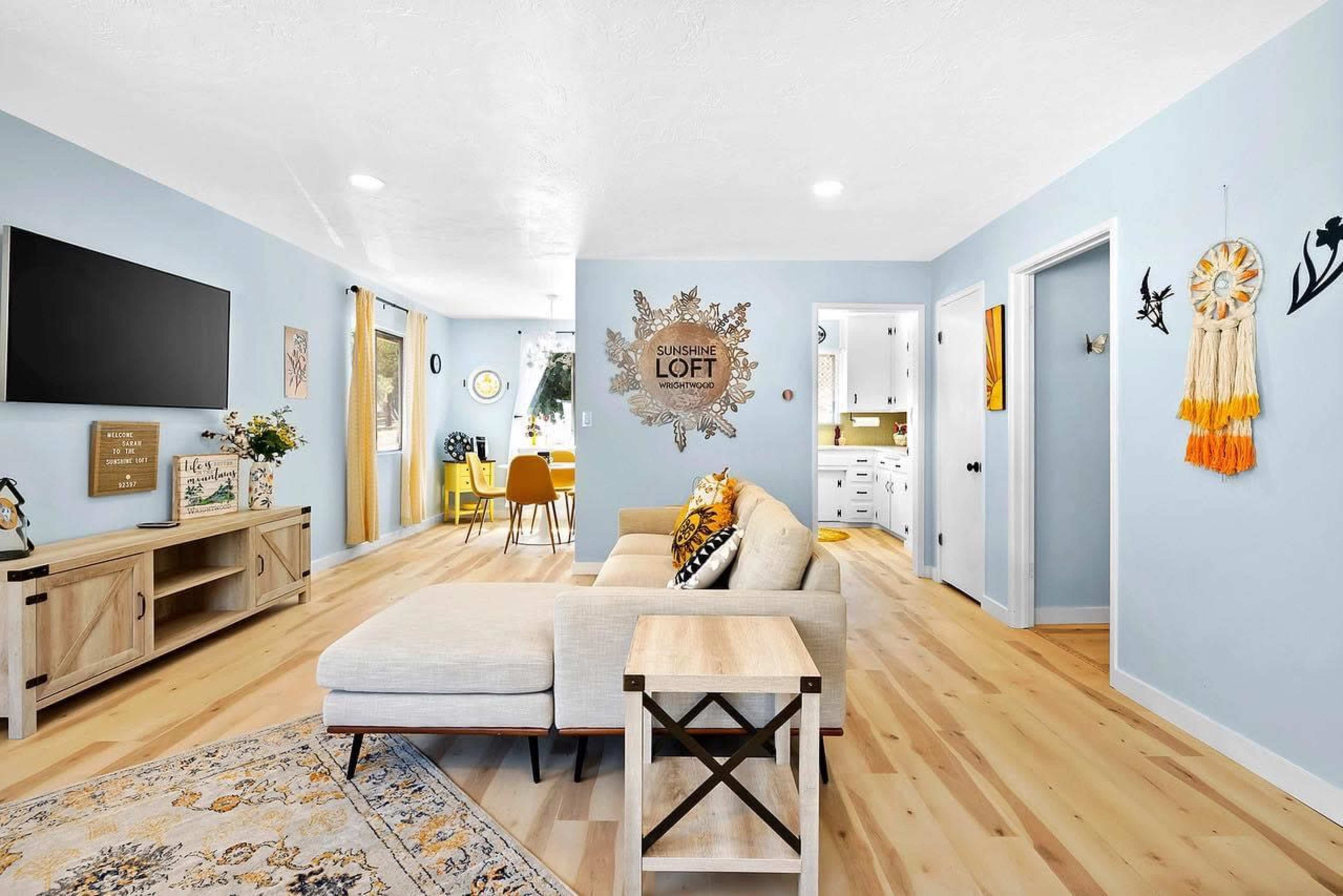 A light blue living room featuring a sectional sofa, a television mounted on the wall, and a wooden coffee table, with a dining area visible in the background.