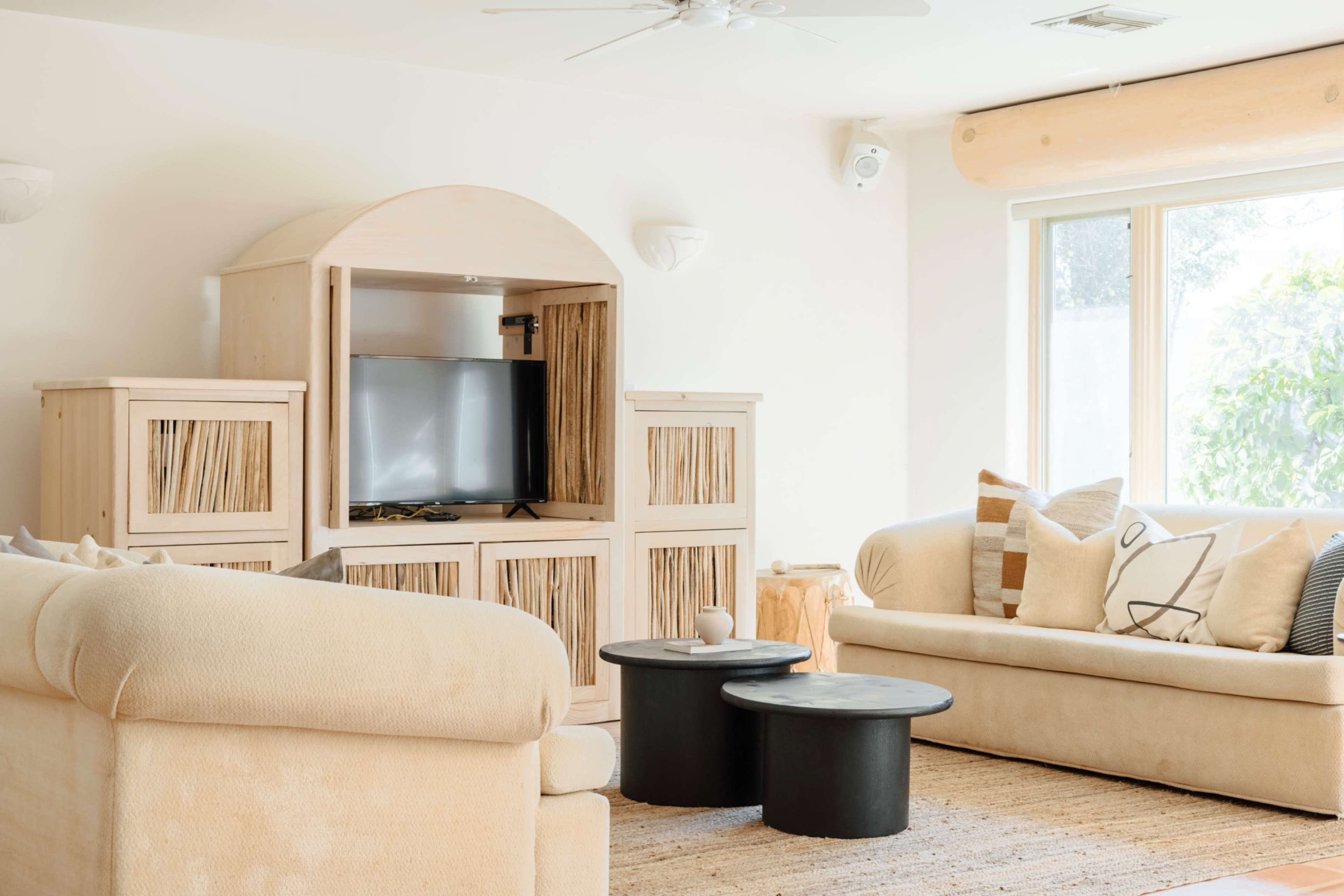 The room features two beige sofas, a wooden entertainment unit with a television, and two black circular coffee tables on a light woven rug.