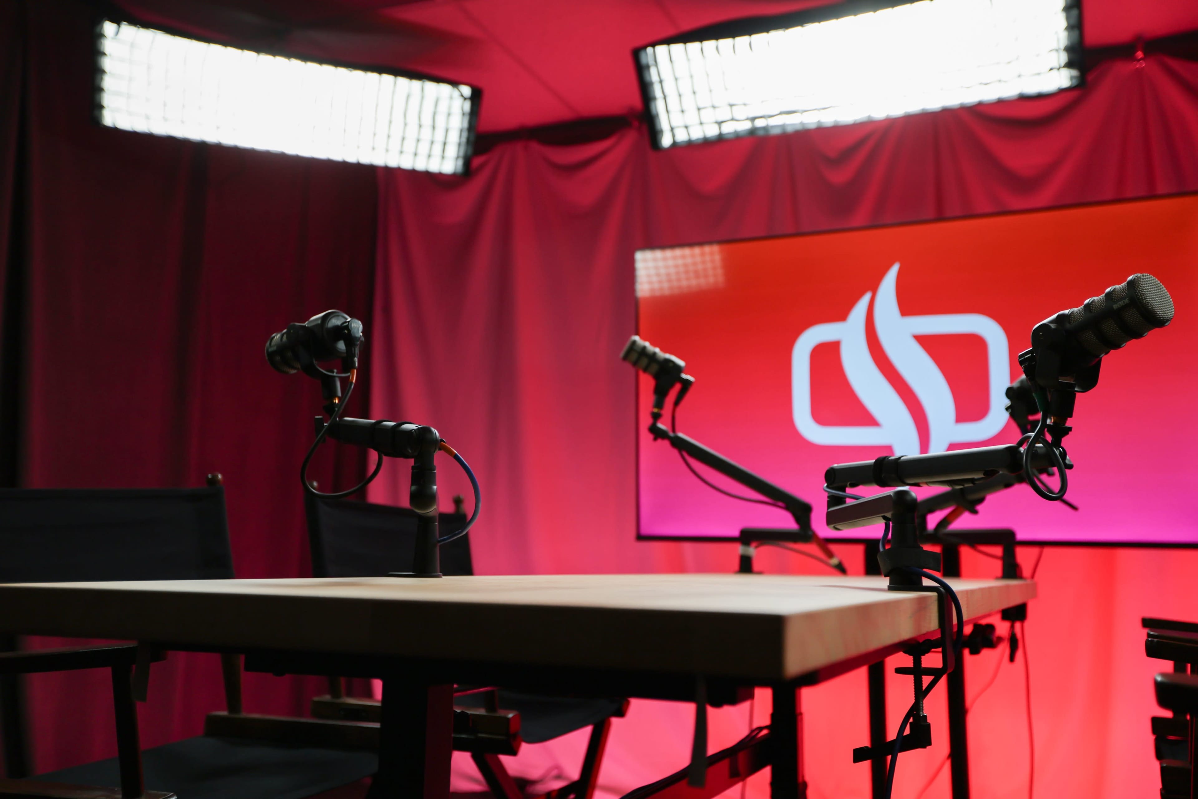 The image shows a podcast or interview setup with a wooden table and two microphone stands facing a screen with a logo against a red backdrop.