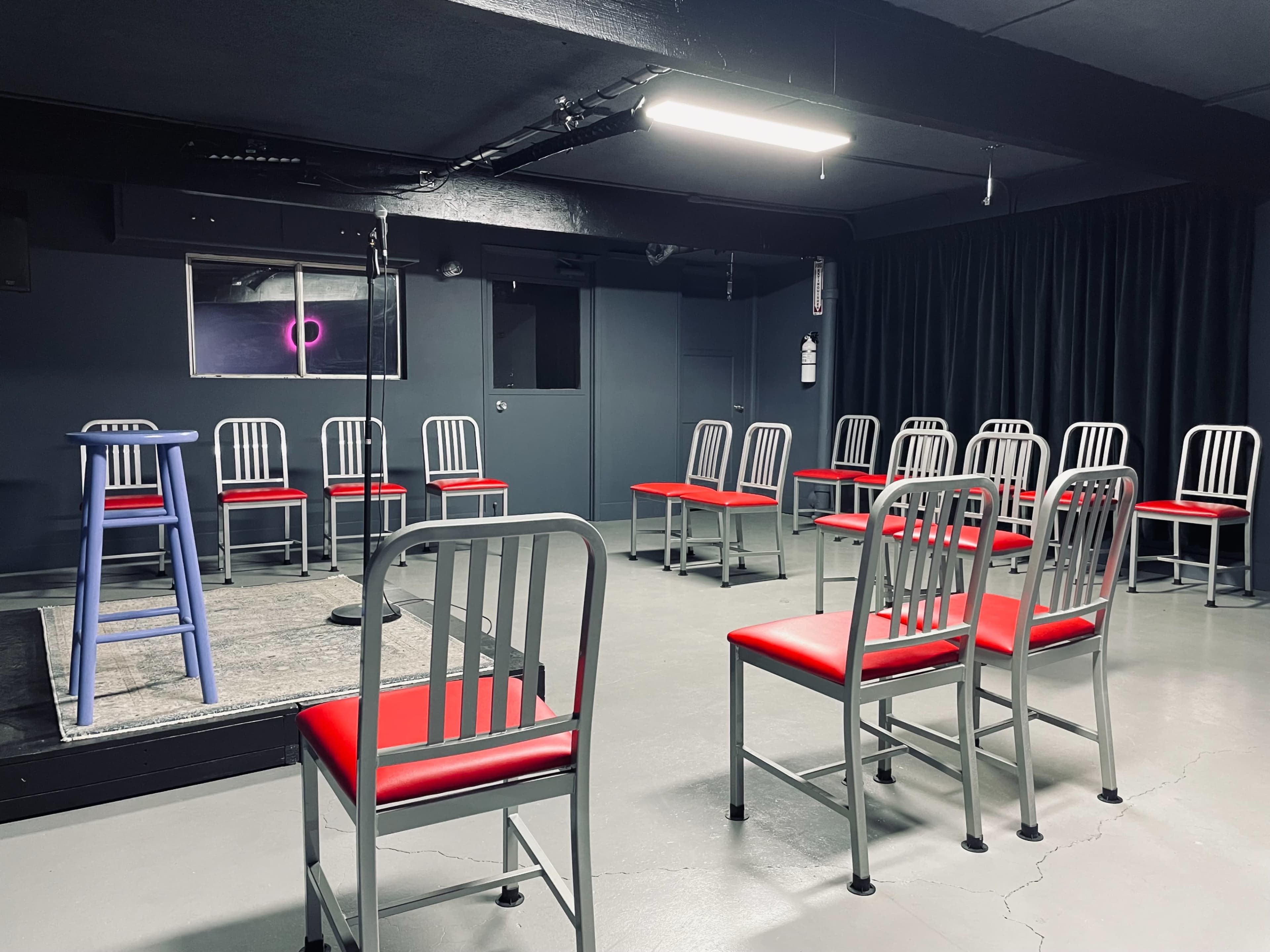 A small performance space features a blue stool and a microphone in front of a circle of red and silver chairs.