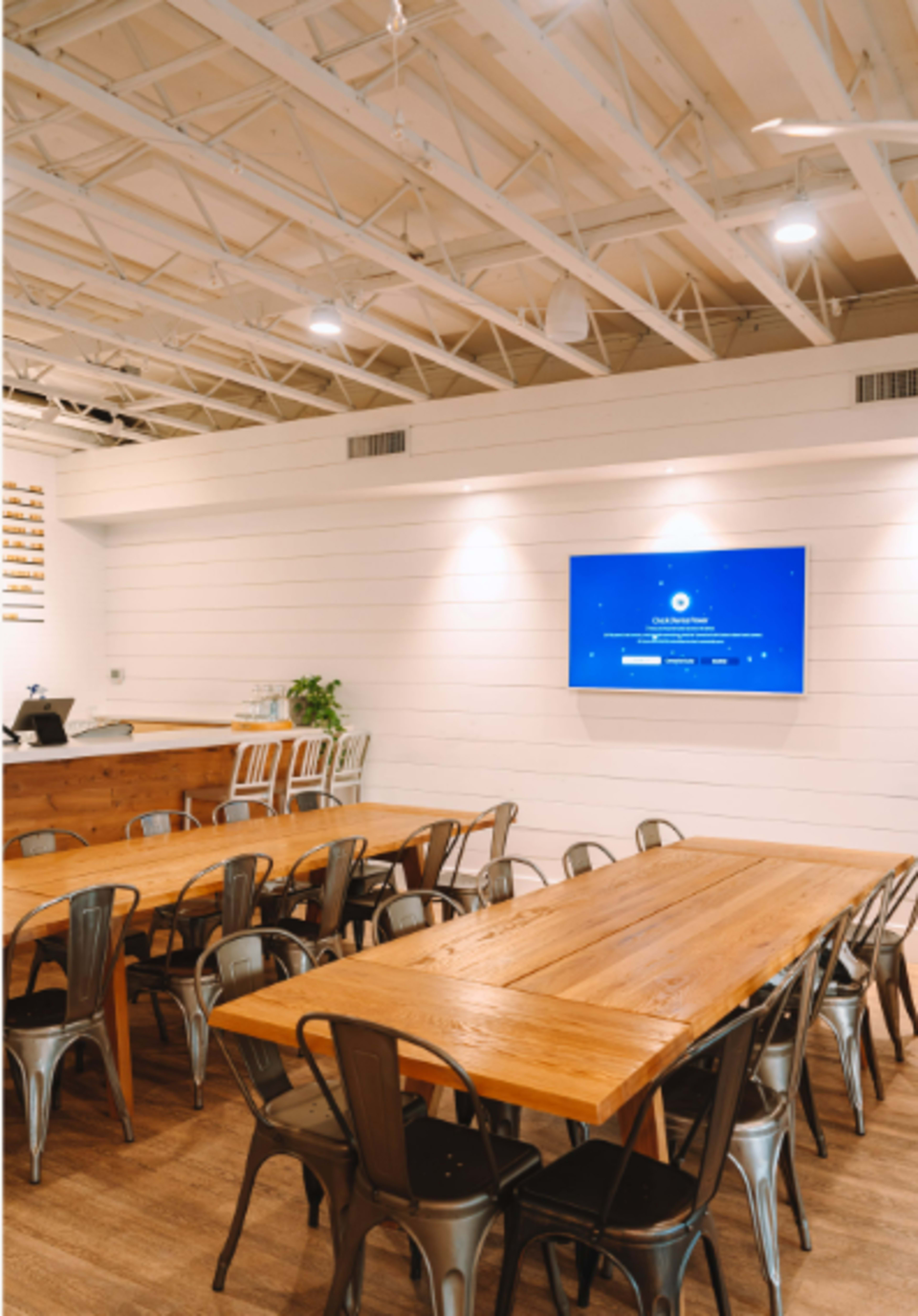 A spacious meeting room features long communal tables with metal chairs and a wall-mounted screen.