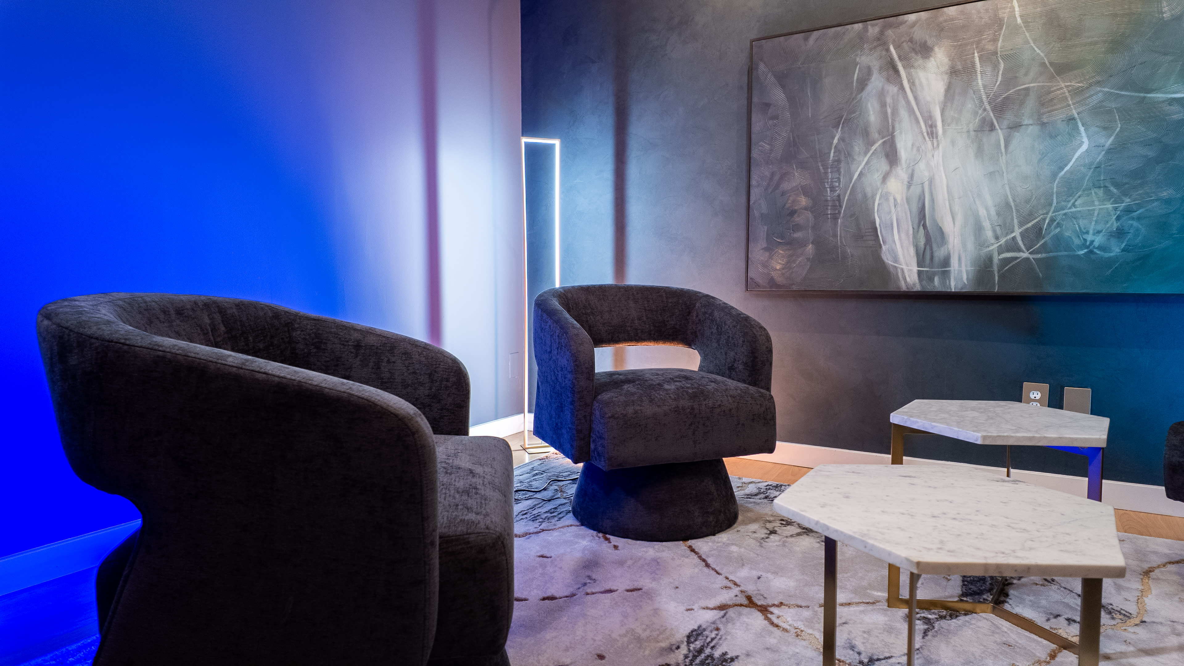 The image shows a modern interior space featuring two dark upholstered chairs, a marble-top coffee table, and an abstract painting on the wall, illuminated by blue light.