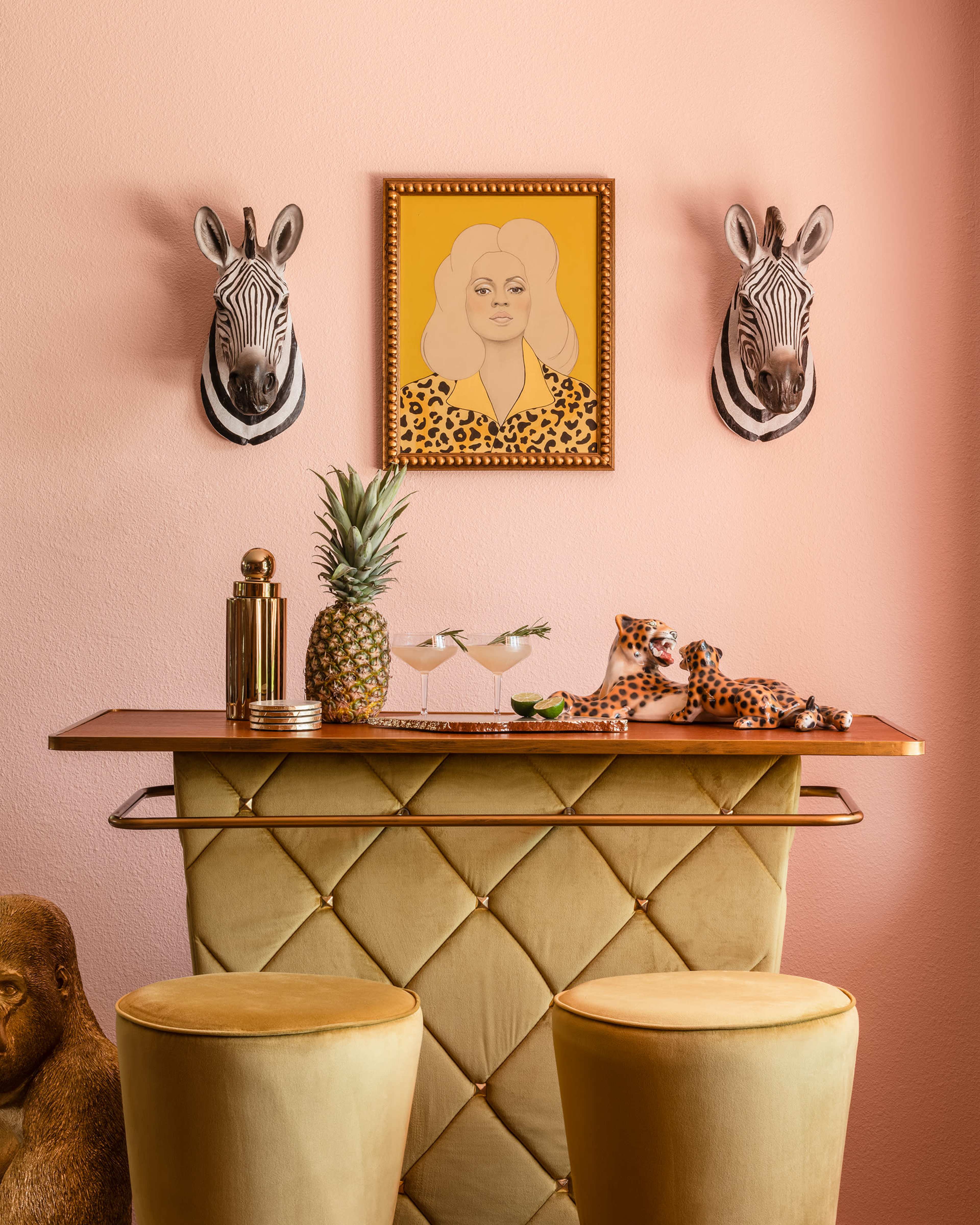 A pink wall with a bar decorated with two zebra head mountings, a portrait of a woman, a pineapple, and two leopard figurines, complemented by two round, yellow stools.