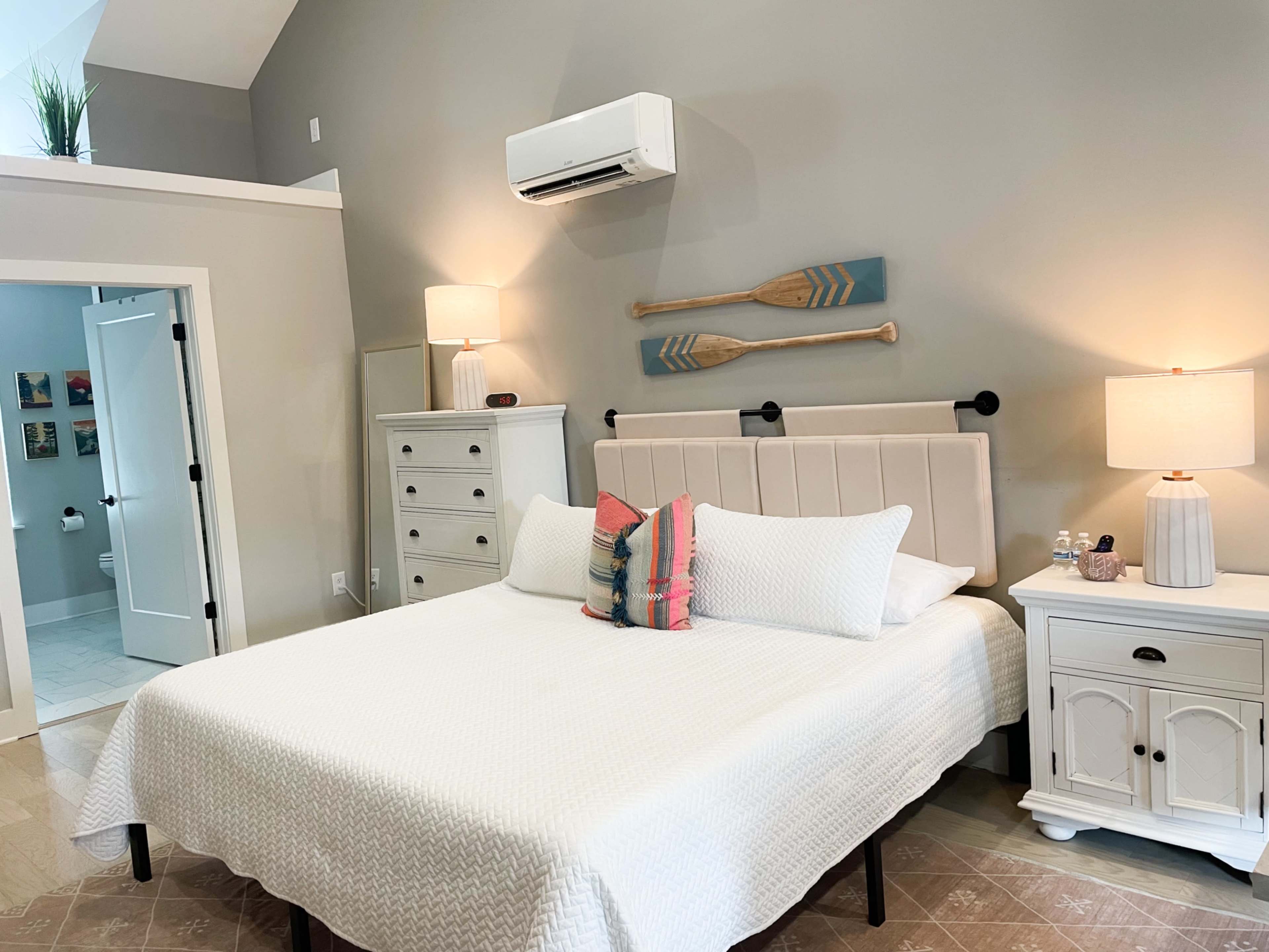 The image shows a neatly arranged bedroom with a white bedspread, nightstands with lamps, a decorative paddles wall art, and light gray walls.