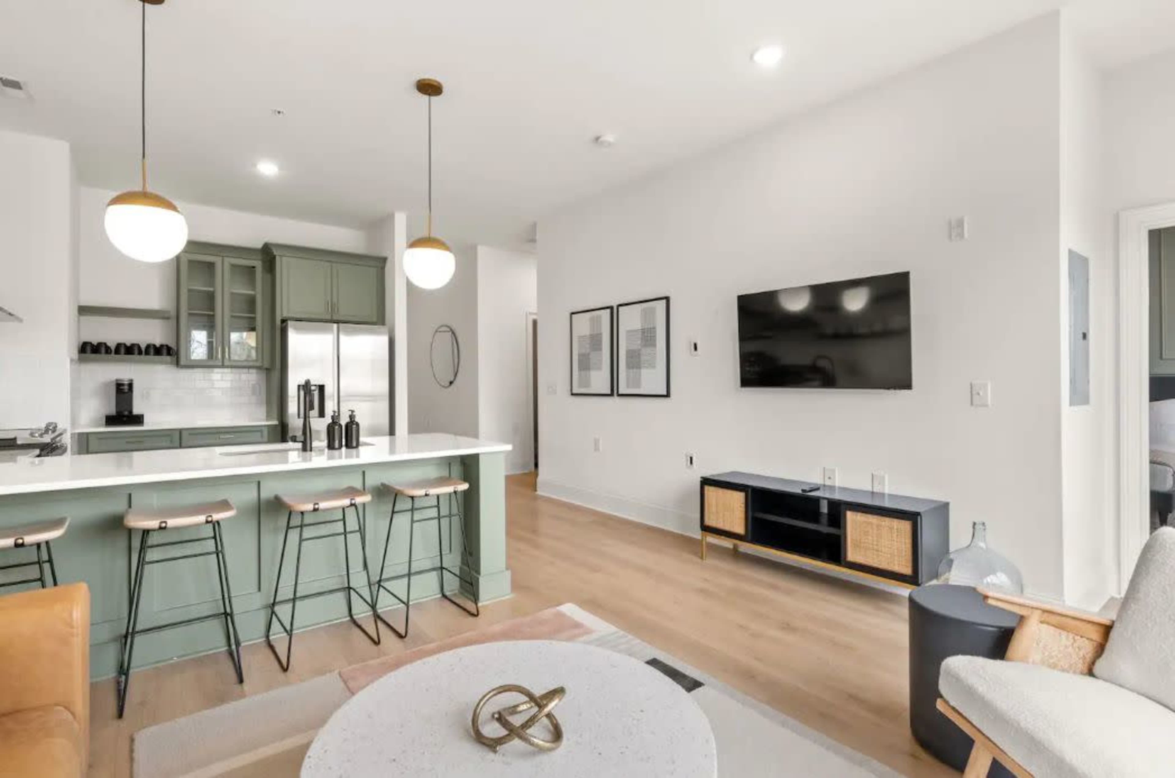A modern kitchen and living area featuring a breakfast bar with three stools, a large television mounted on the wall, and a light-colored sofa.