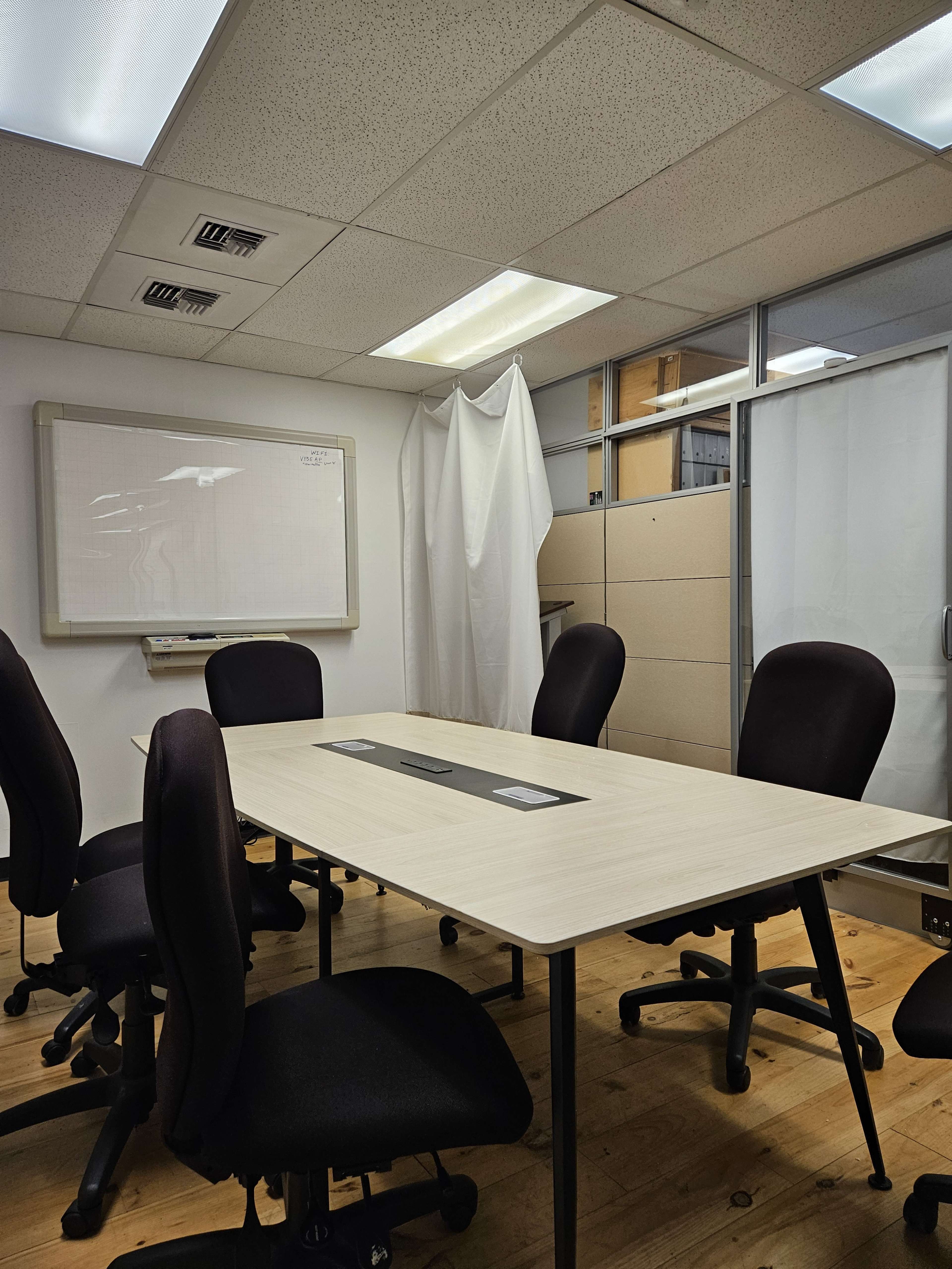 An empty conference room features a central table surrounded by black chairs, with a whiteboard and partition in the background.