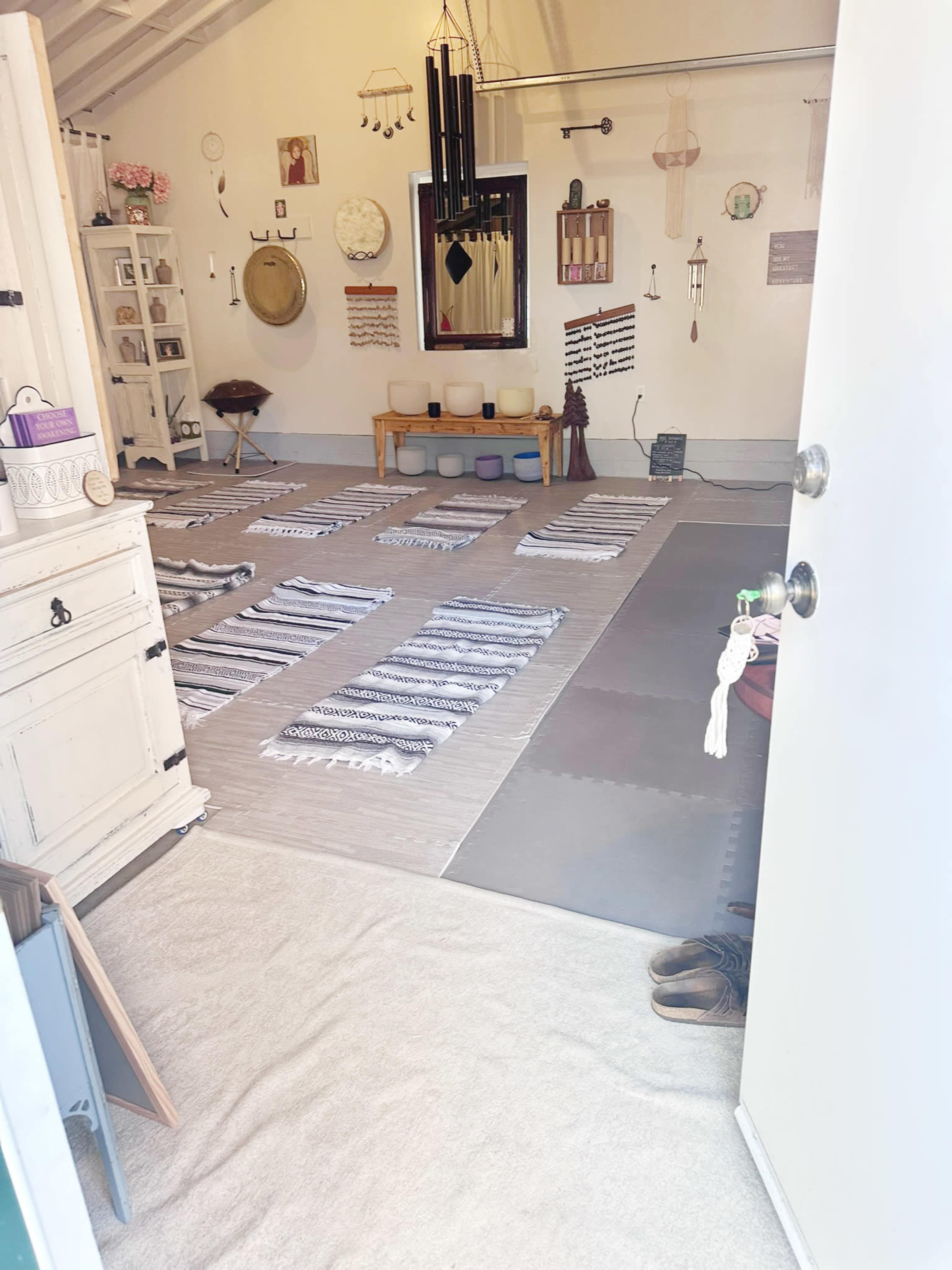 The image shows a yoga or meditation studio with mats arranged on a soft floor, various decorations on the walls, and a welcoming entrance.