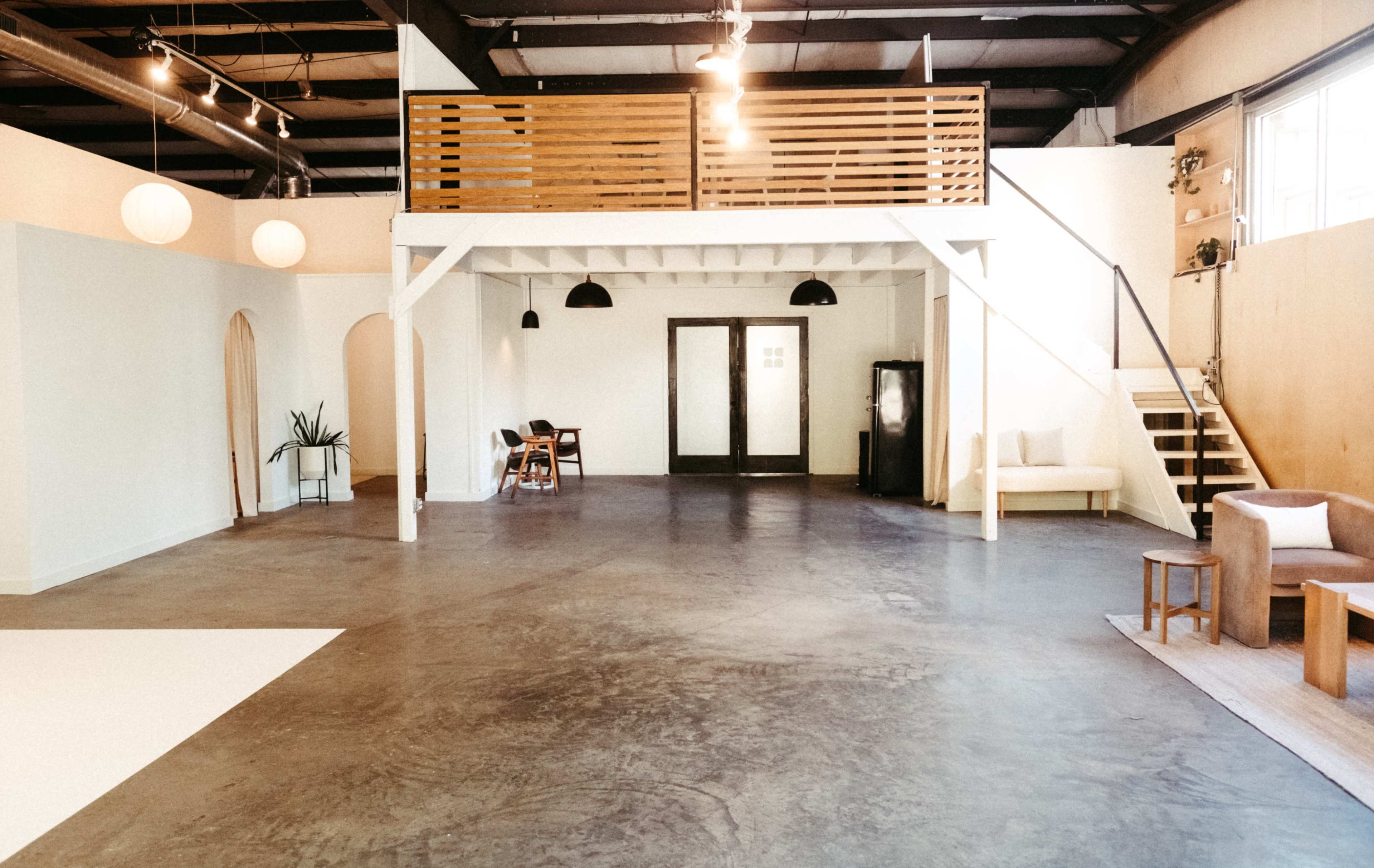 The image shows a spacious, minimalist interior with concrete flooring, a wooden mezzanine, and various seating options, including a sofa and chairs.