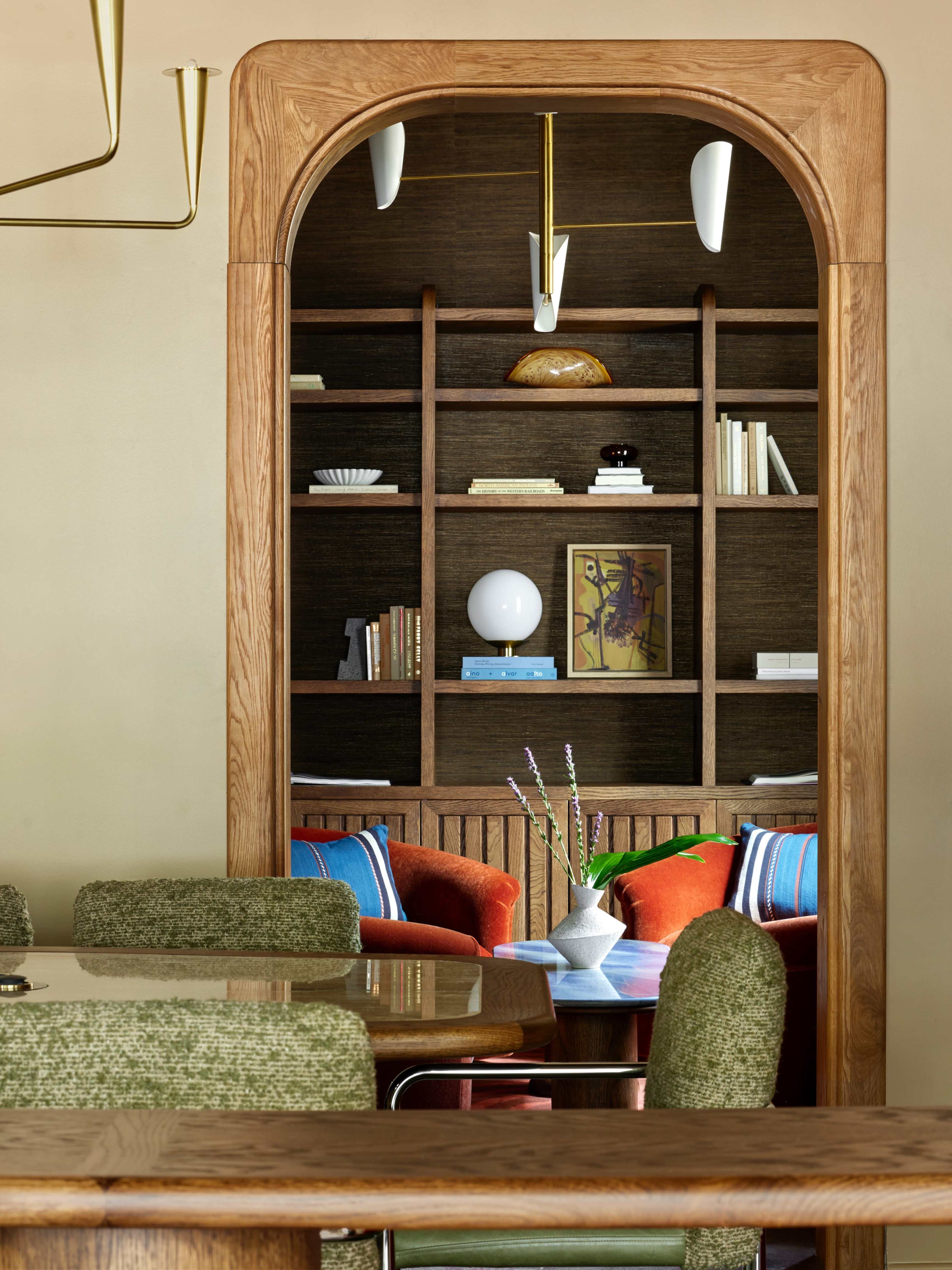A wooden archway frames a neatly arranged bookshelf filled with books and decorative items, with a cozy seating area visible behind it.