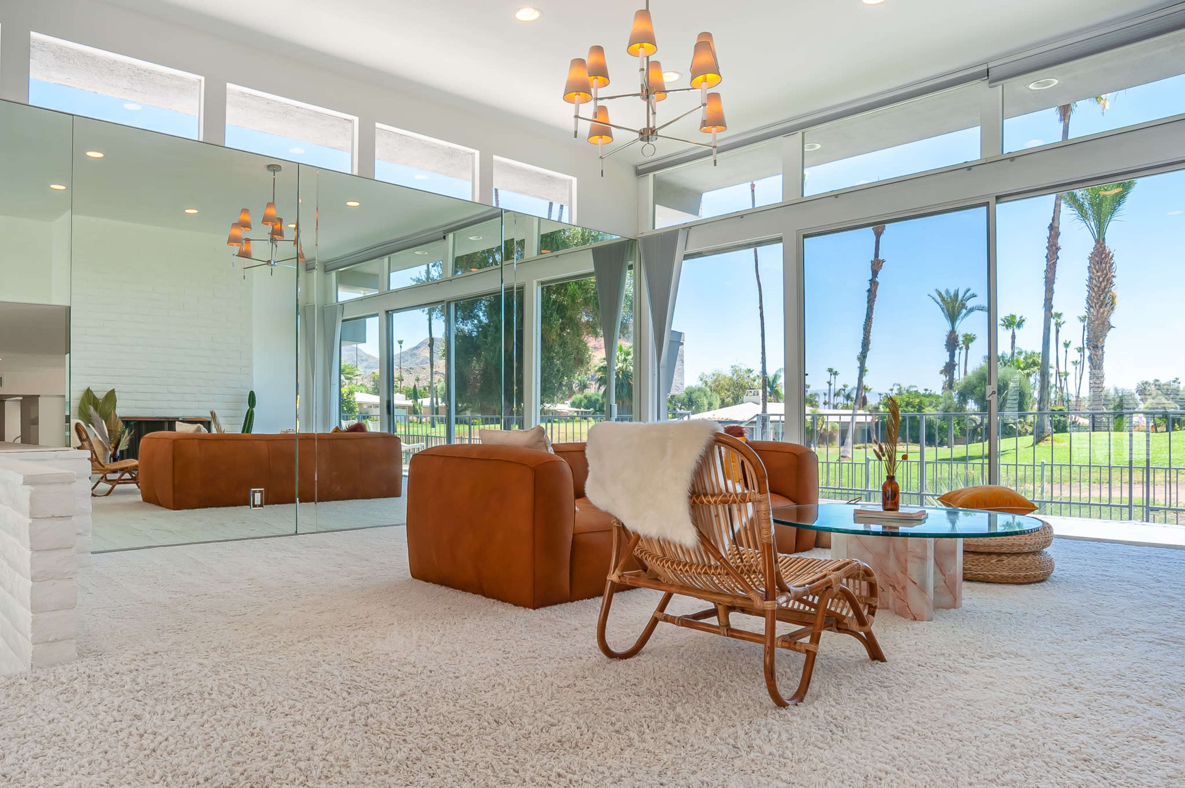 The scene shows a bright, modern living room with large glass windows, reflecting walls, and stylish furniture, including a leather couch and a rattan chair.