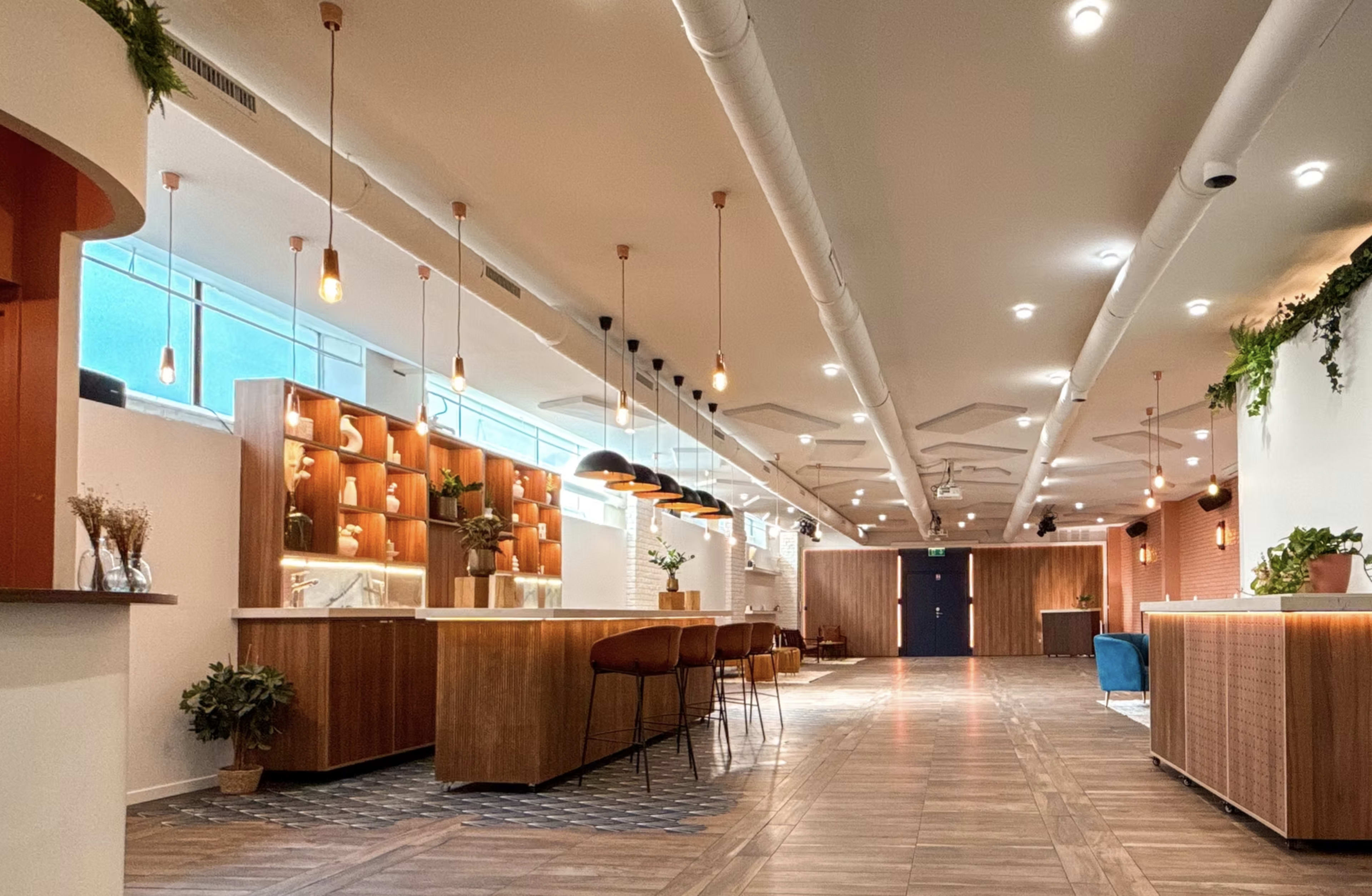 The image shows a modern interior space with wooden accents, featuring a long seating area, a bar-like counter, and overhead pendant lighting.