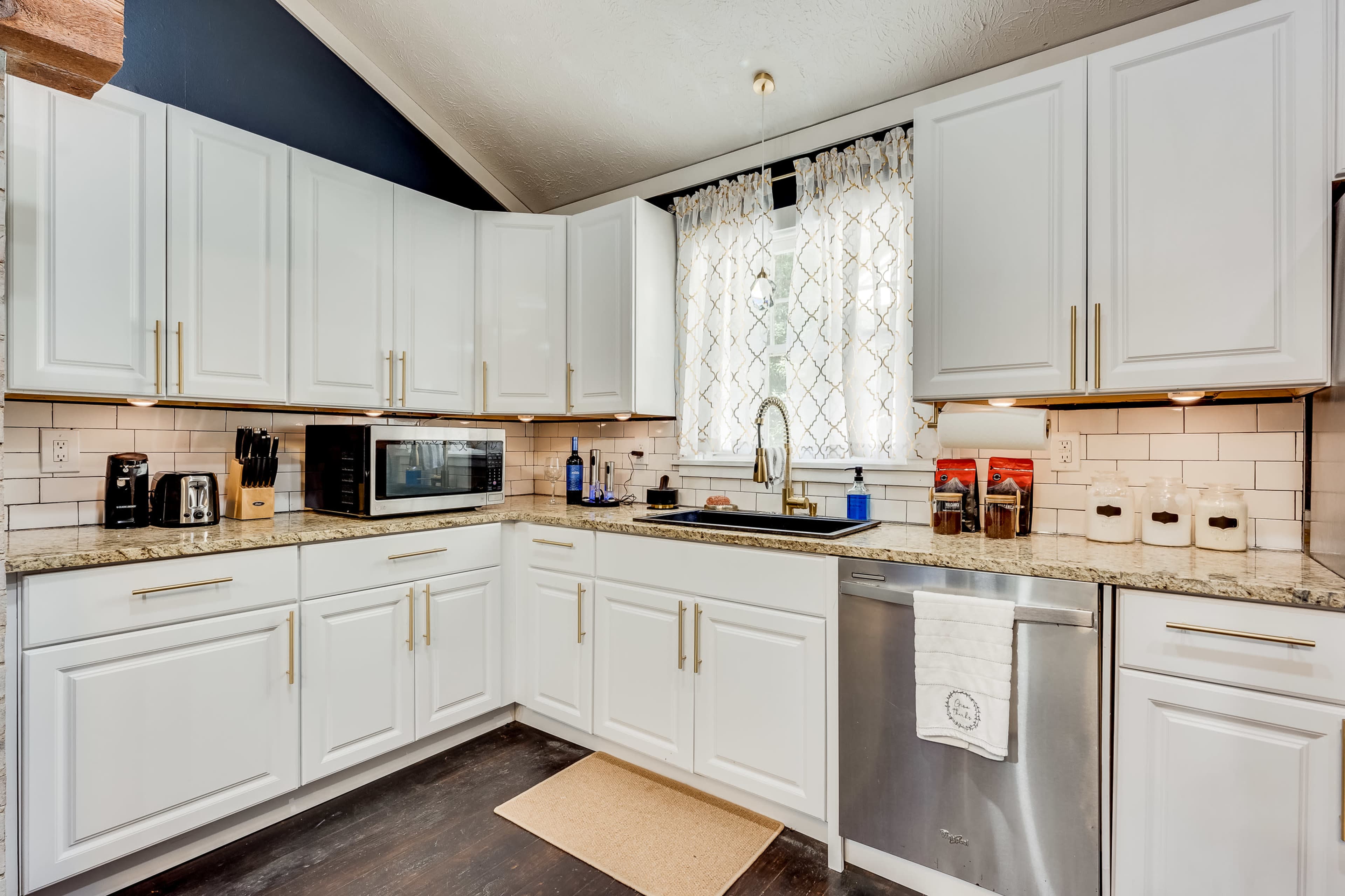 A modern kitchen with white cabinets, a stainless steel dishwasher, a microwave, and a window with a patterned curtain above the sink.
