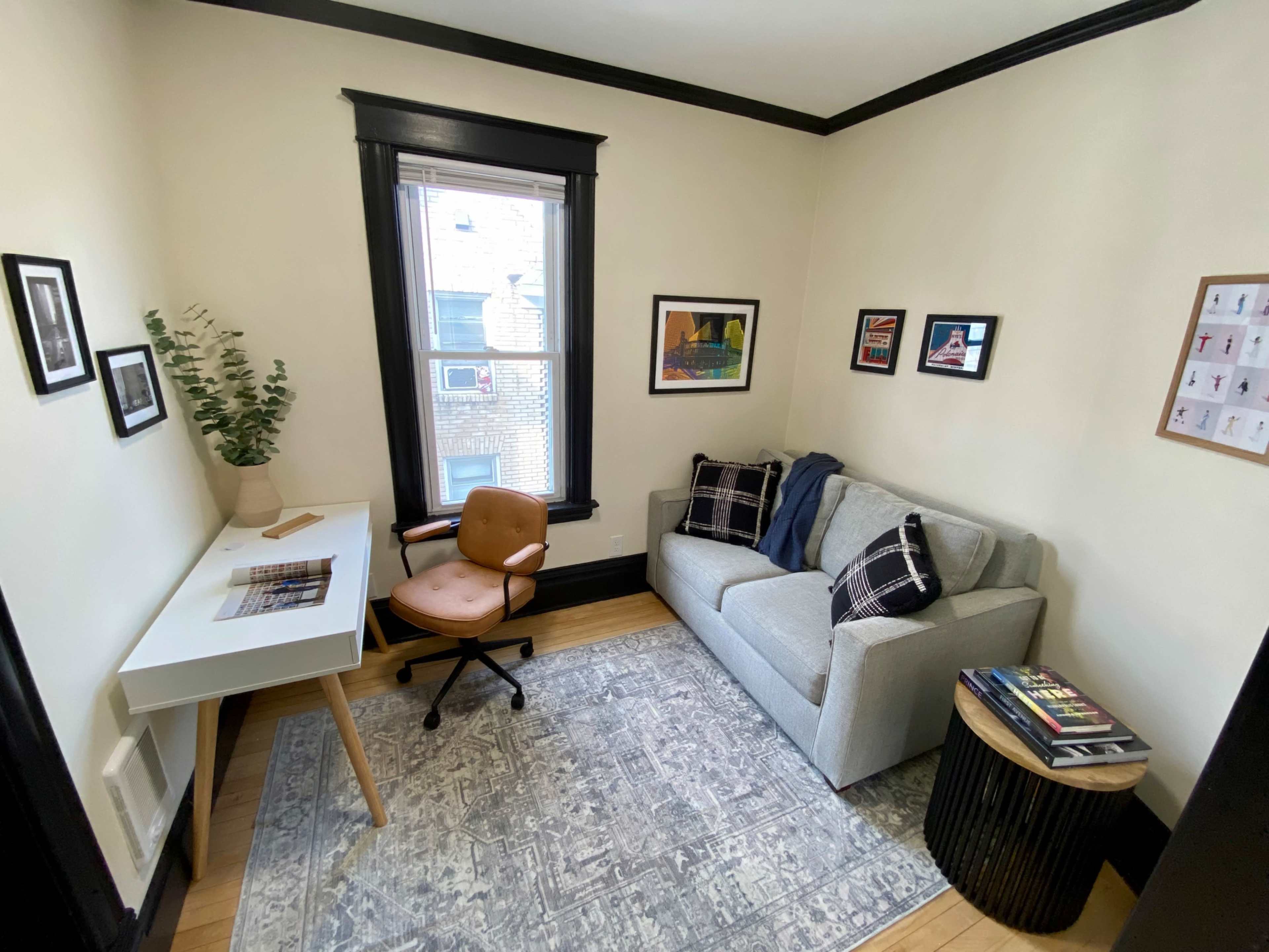 A small office space features a desk with a chair, a couch, and framed artworks on the walls.