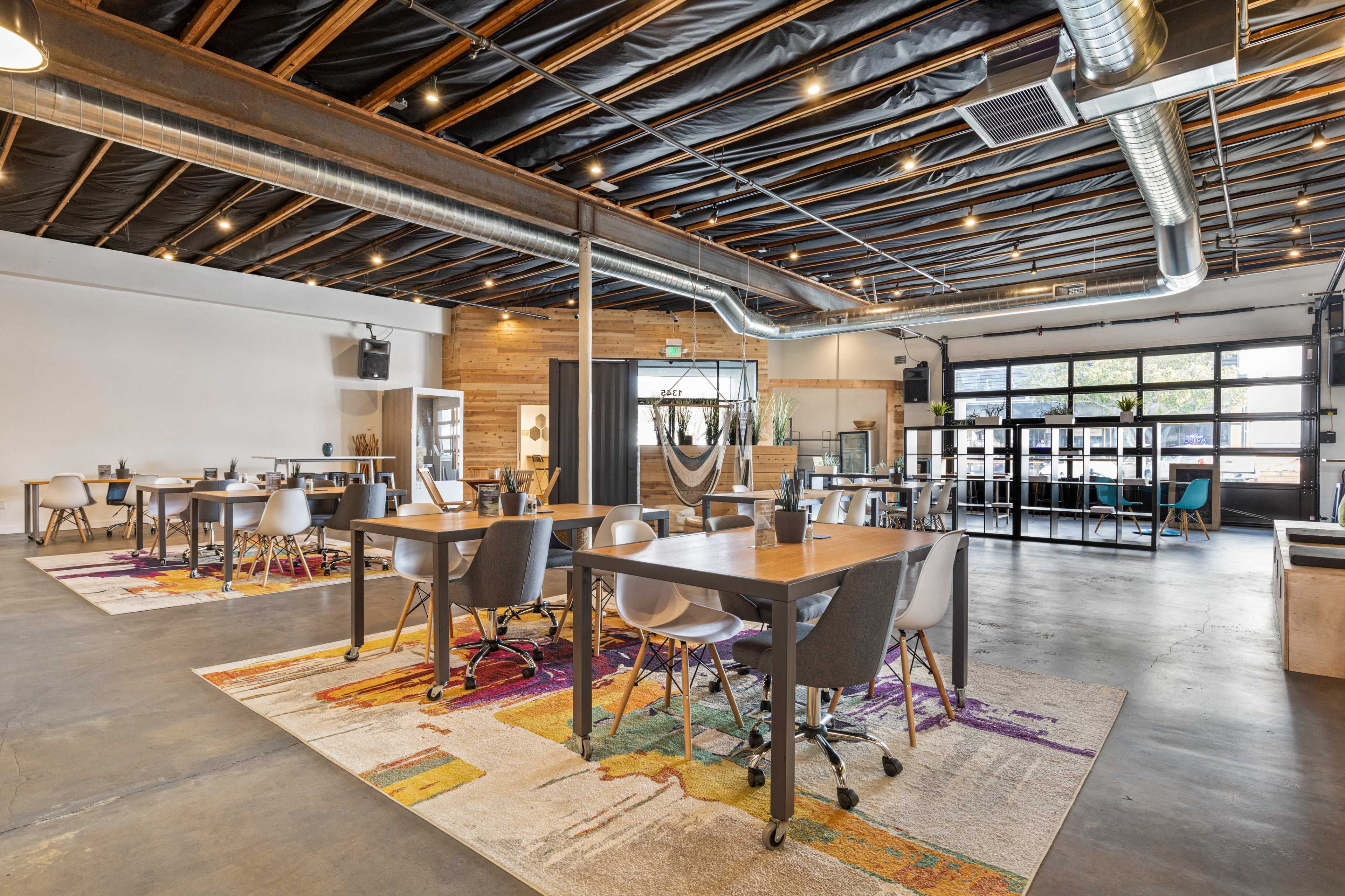 The image shows a modern indoor space featuring several wooden tables and chairs arranged on a colorful carpet, with industrial-style lighting and large windows.