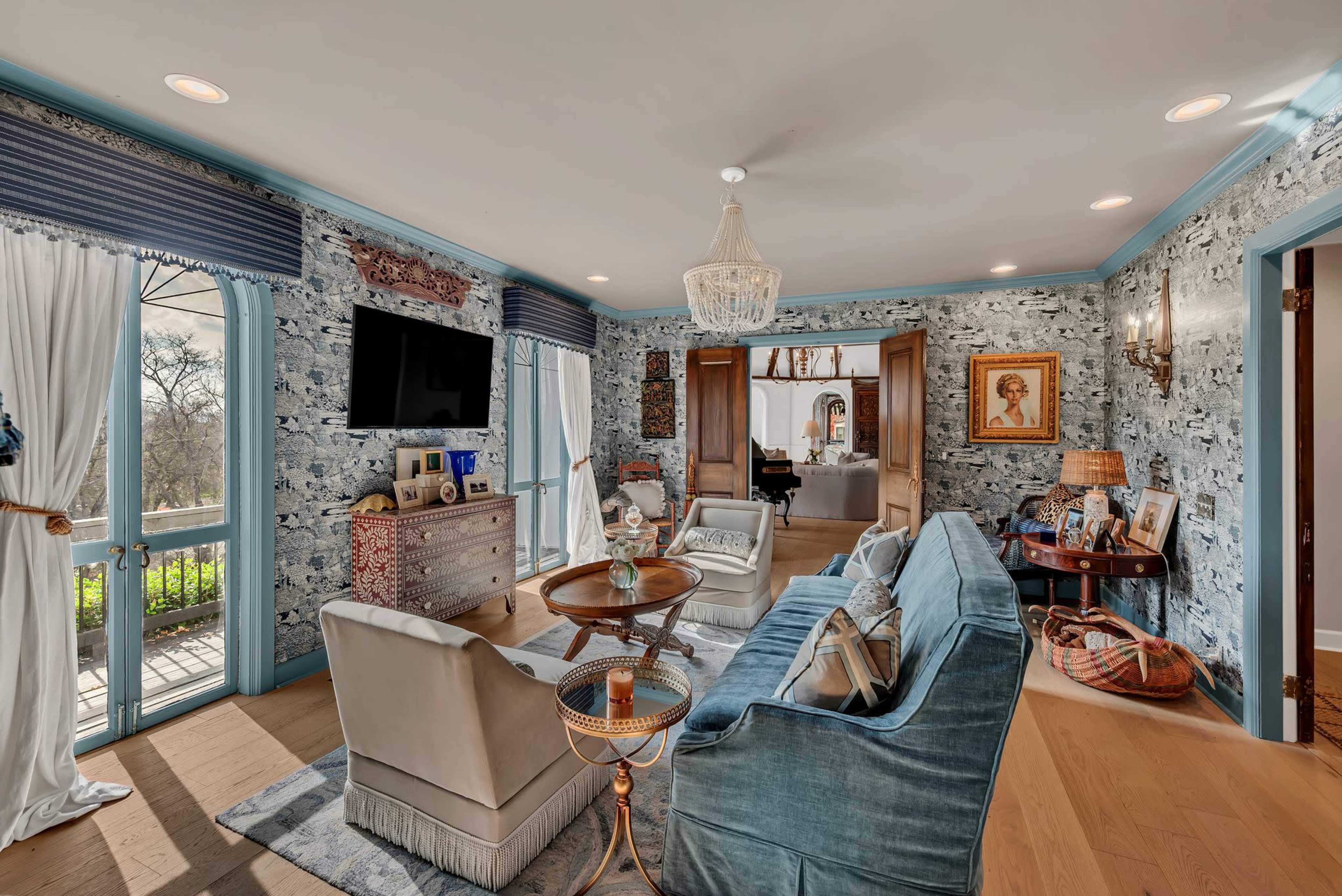 A bright living room features a blue and white wallpaper design, a comfortable blue sofa, two cream-colored chairs, and a decorative chandelier.