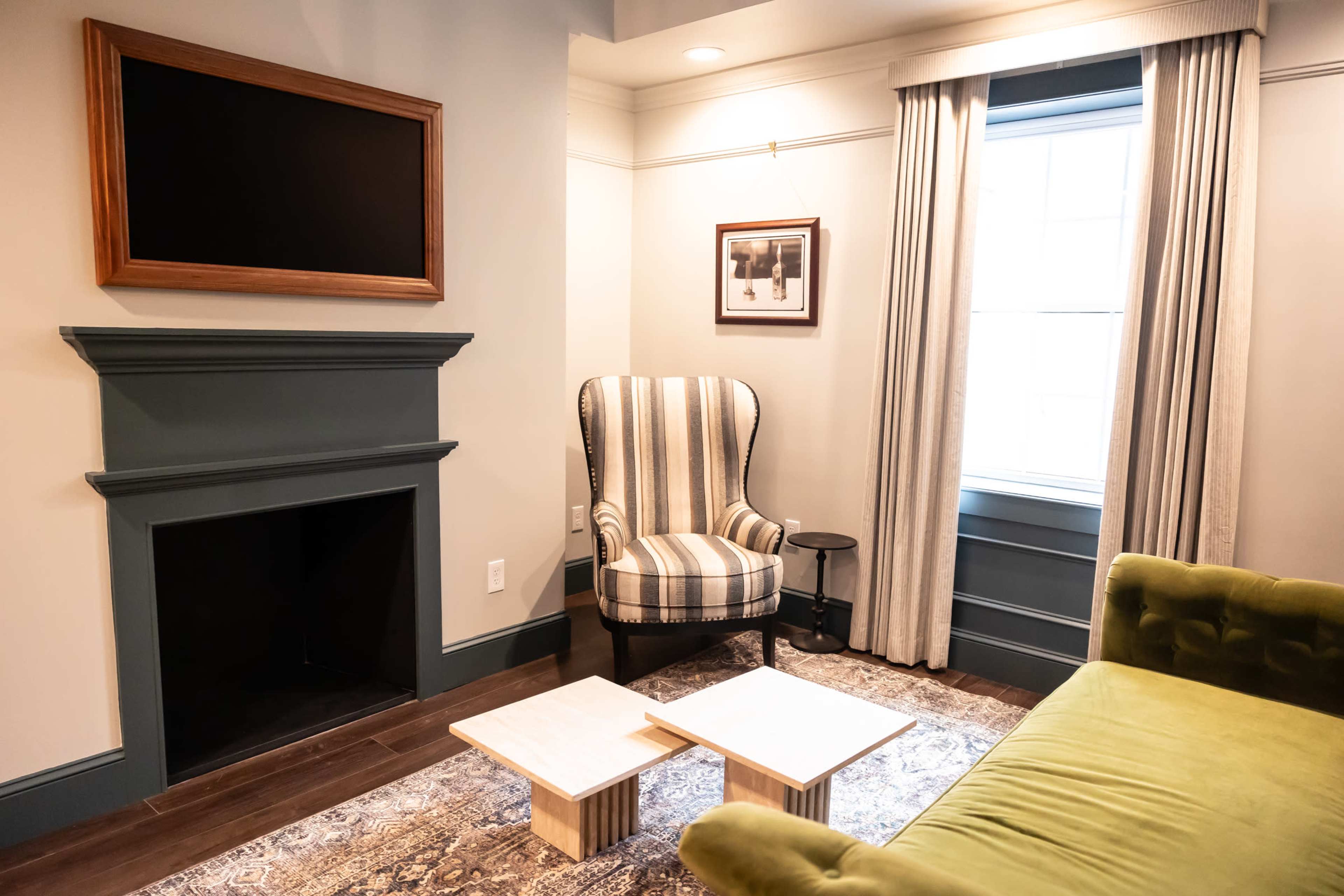 A cozy living room features a striped armchair, a green sofa, and two small tables on a patterned rug near a fireplace.