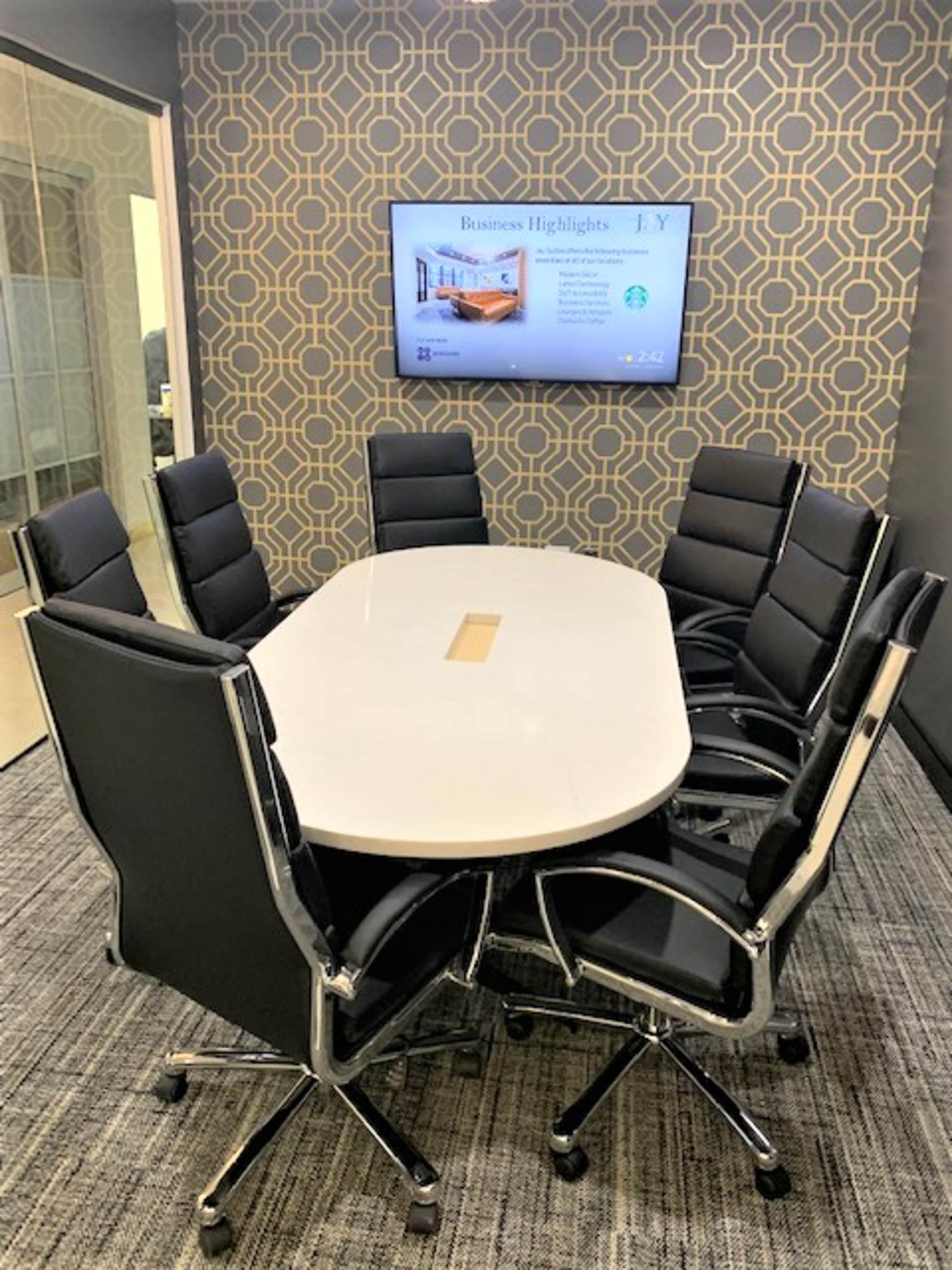 A modern conference room features a large oval table surrounded by black rolling chairs and a mounted screen displaying business highlights on a geometric patterned wall.