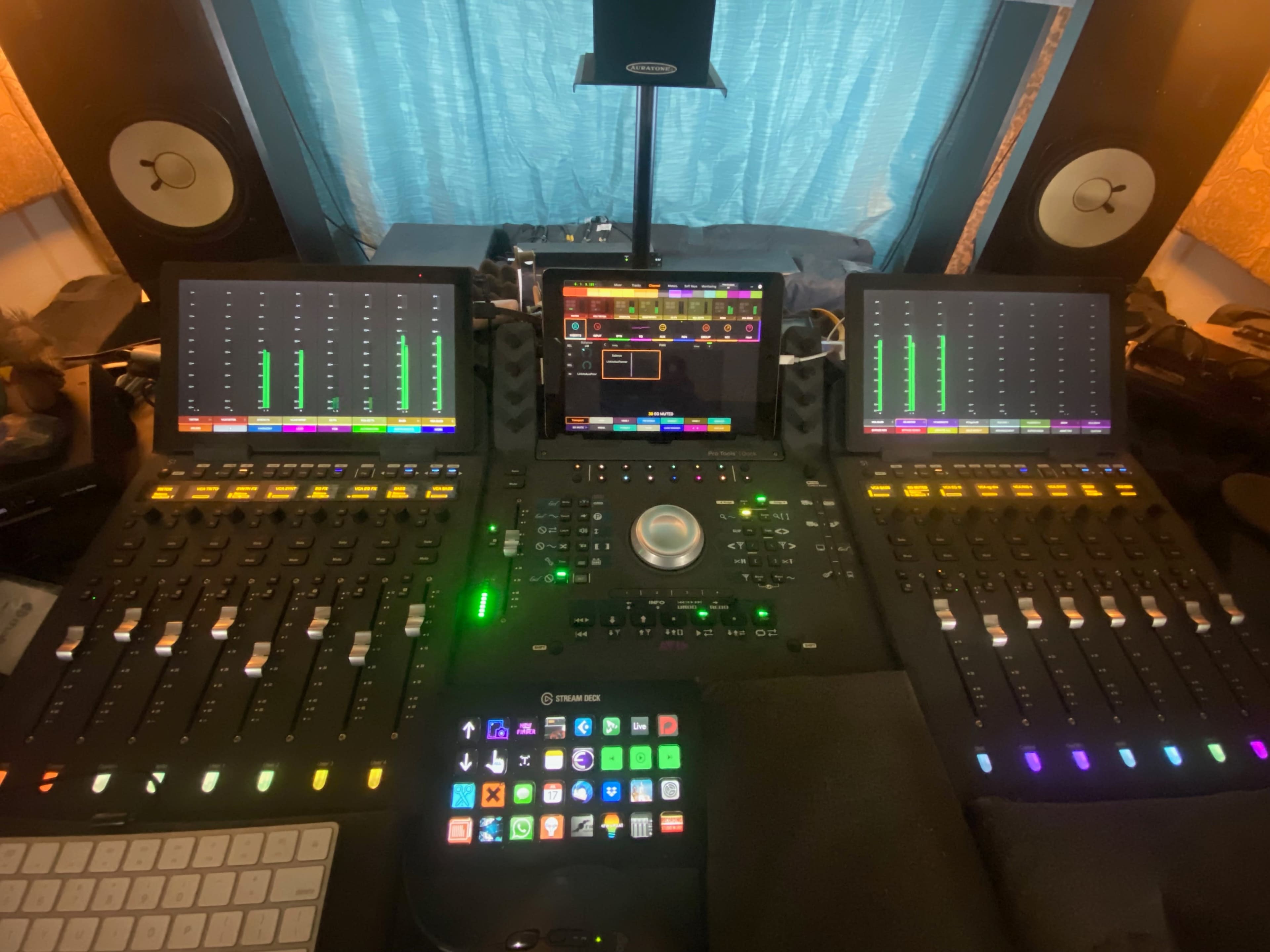 A digital audio workstation setup featuring multiple control surfaces, touchscreen displays, and studio monitors in a room with blue curtains.