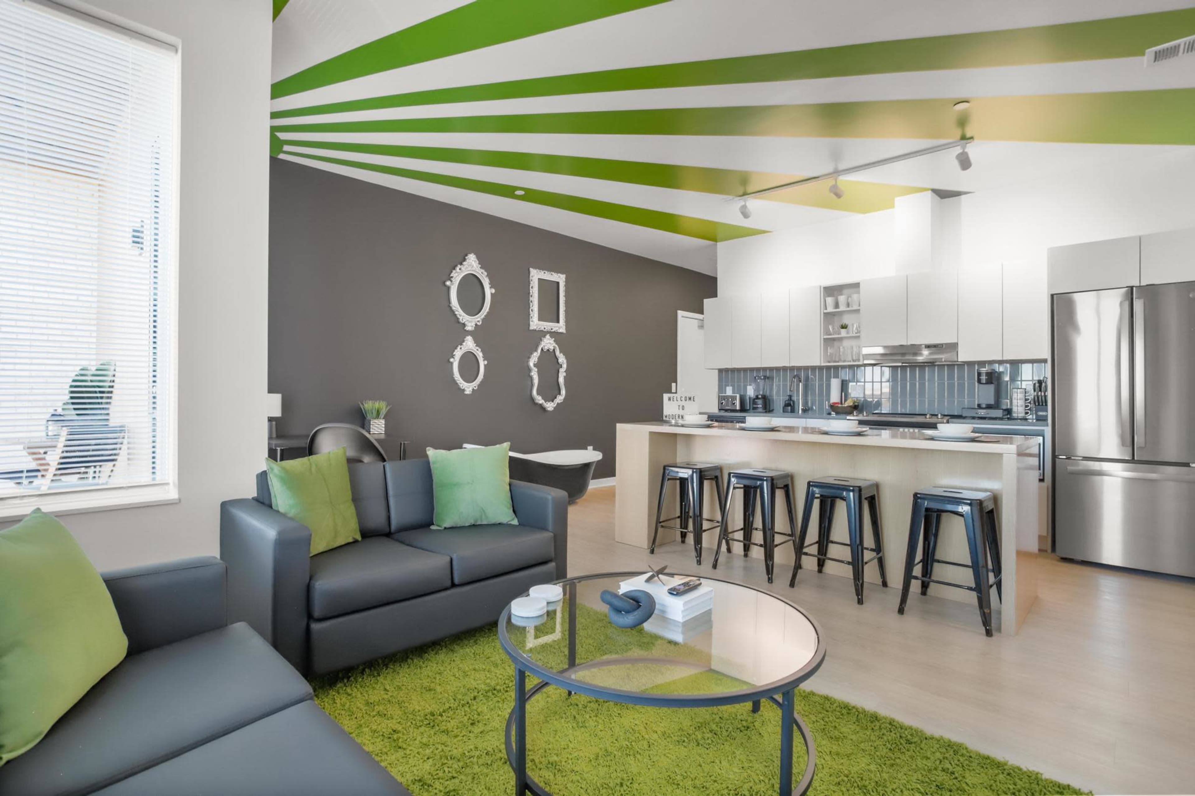 The image shows a modern open-concept living space featuring a kitchen with white cabinetry, a gray accent wall, and green striped ceiling beams, accompanied by a seating area with a black sofa and green cushions.