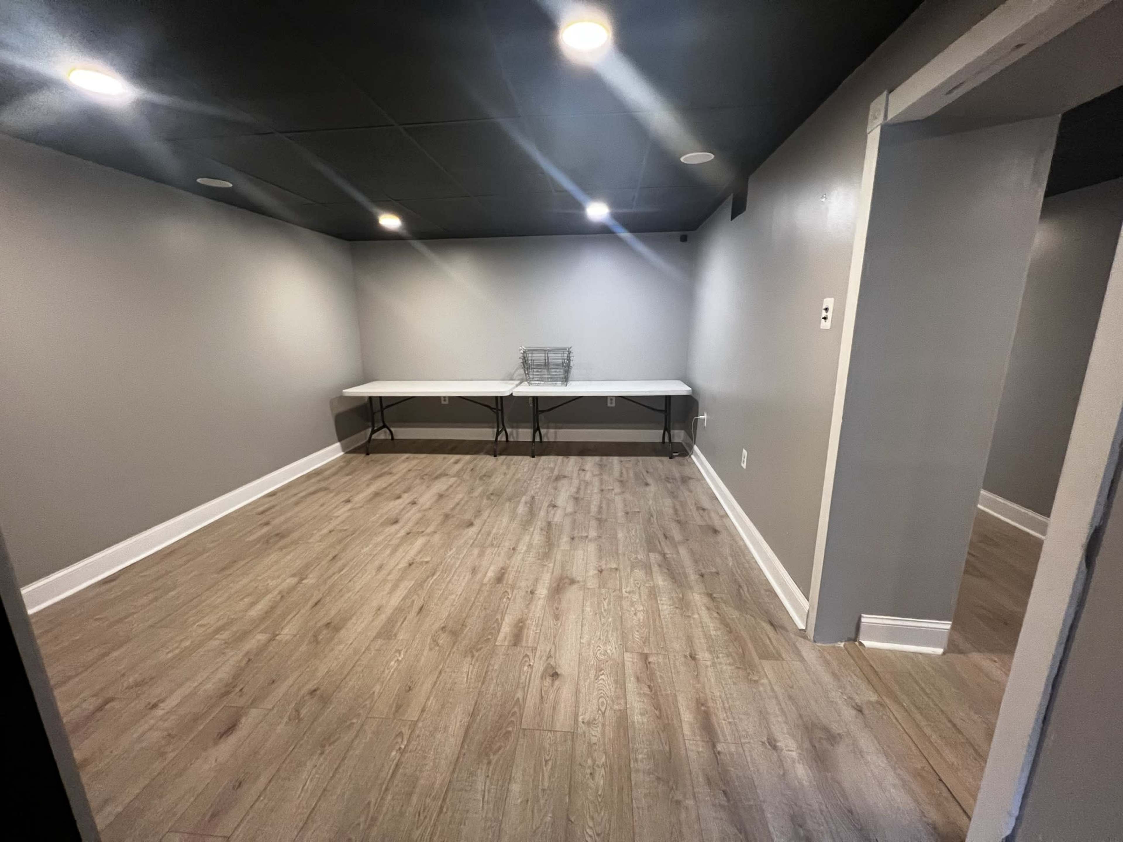 A vacant room with gray walls, wooden flooring, and two folding tables in the center under overhead lighting.