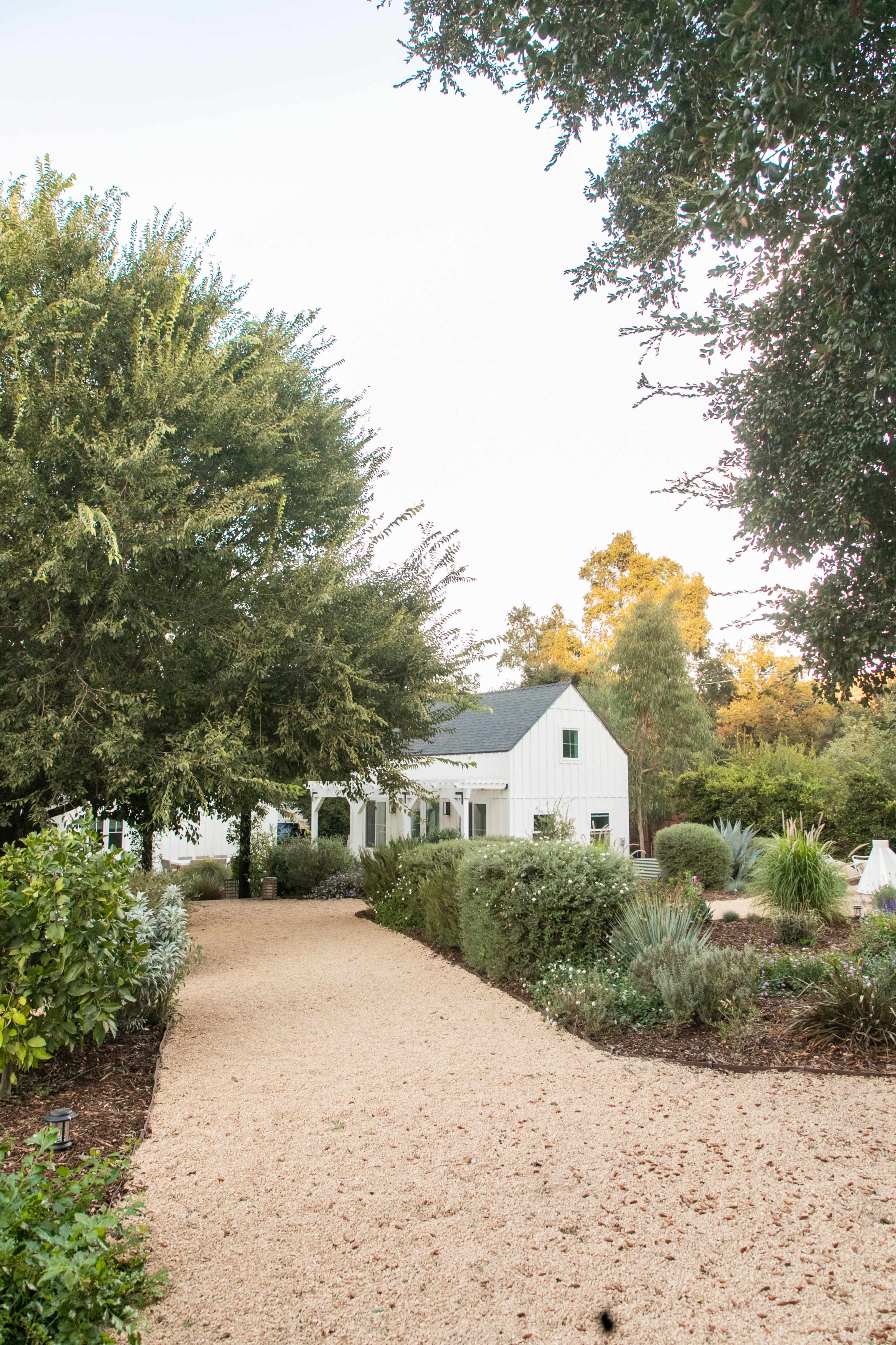 A gravel pathway leads to a white farmhouse surrounded by trees and well-maintained landscaping.