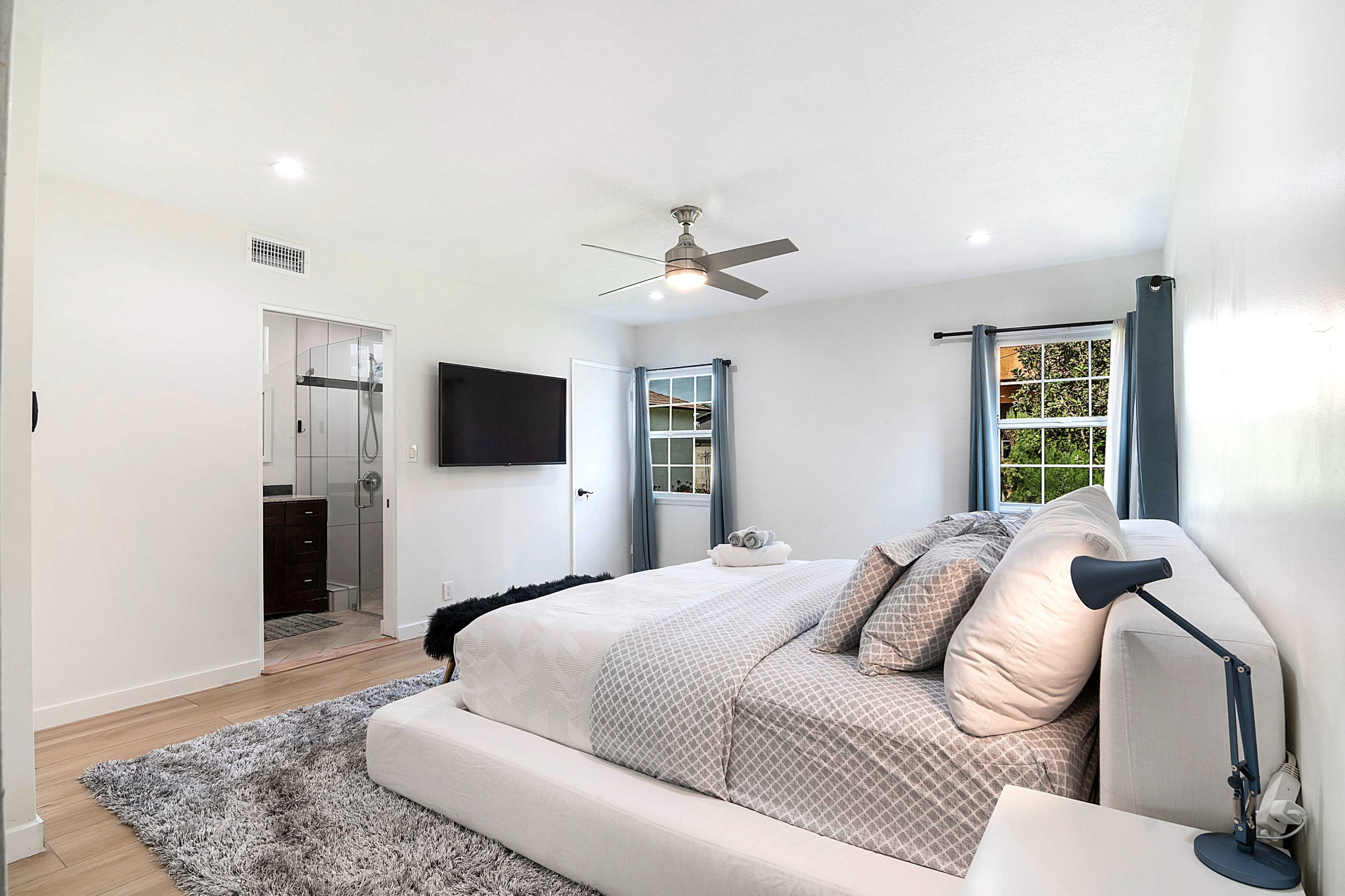 The image shows a modern bedroom with a large bed, a mounted television, and an en-suite bathroom visible through a doorway.