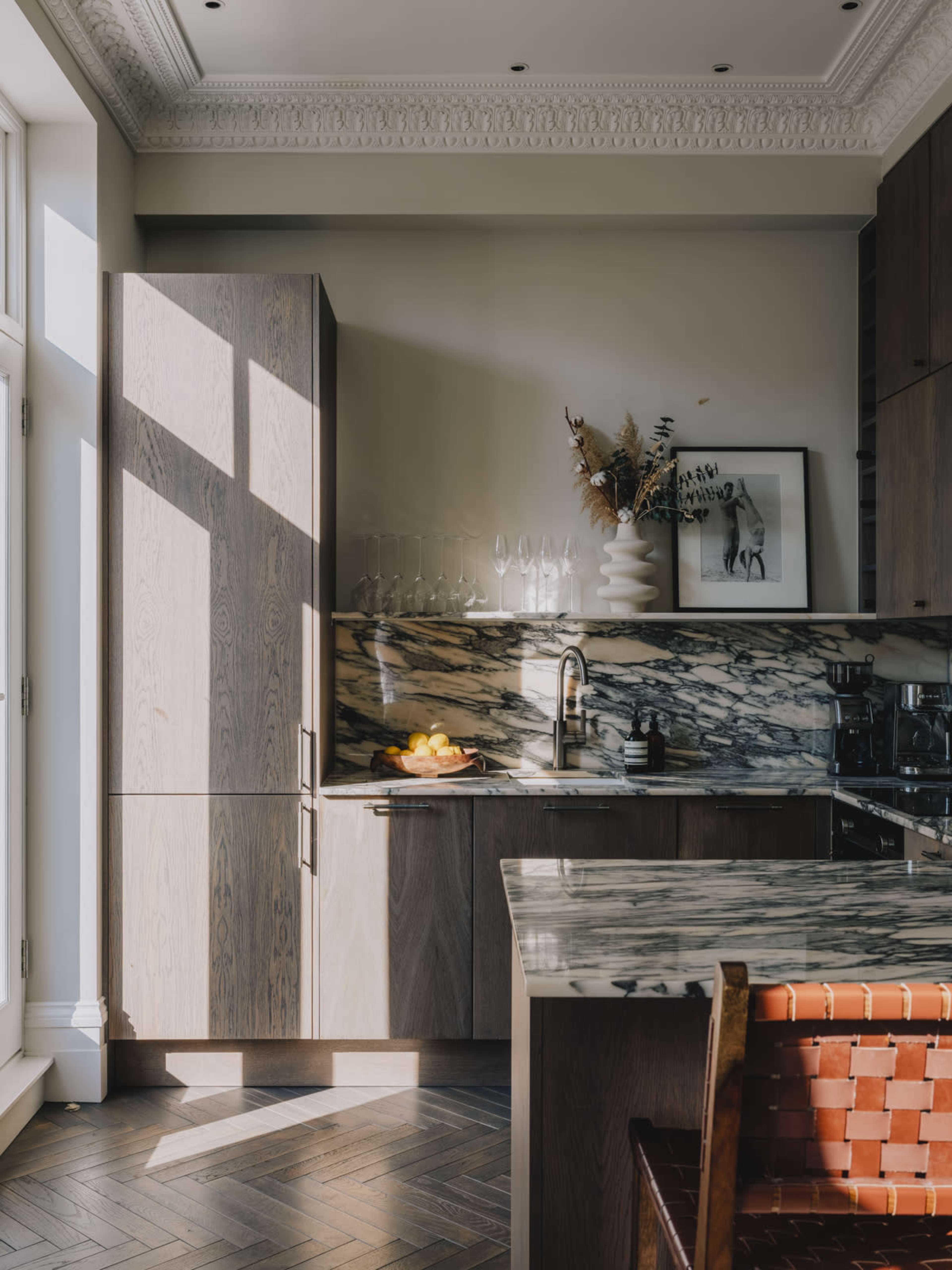 The image depicts a modern kitchen with wooden cabinetry, marble countertops, and large windows allowing natural light to illuminate the space.