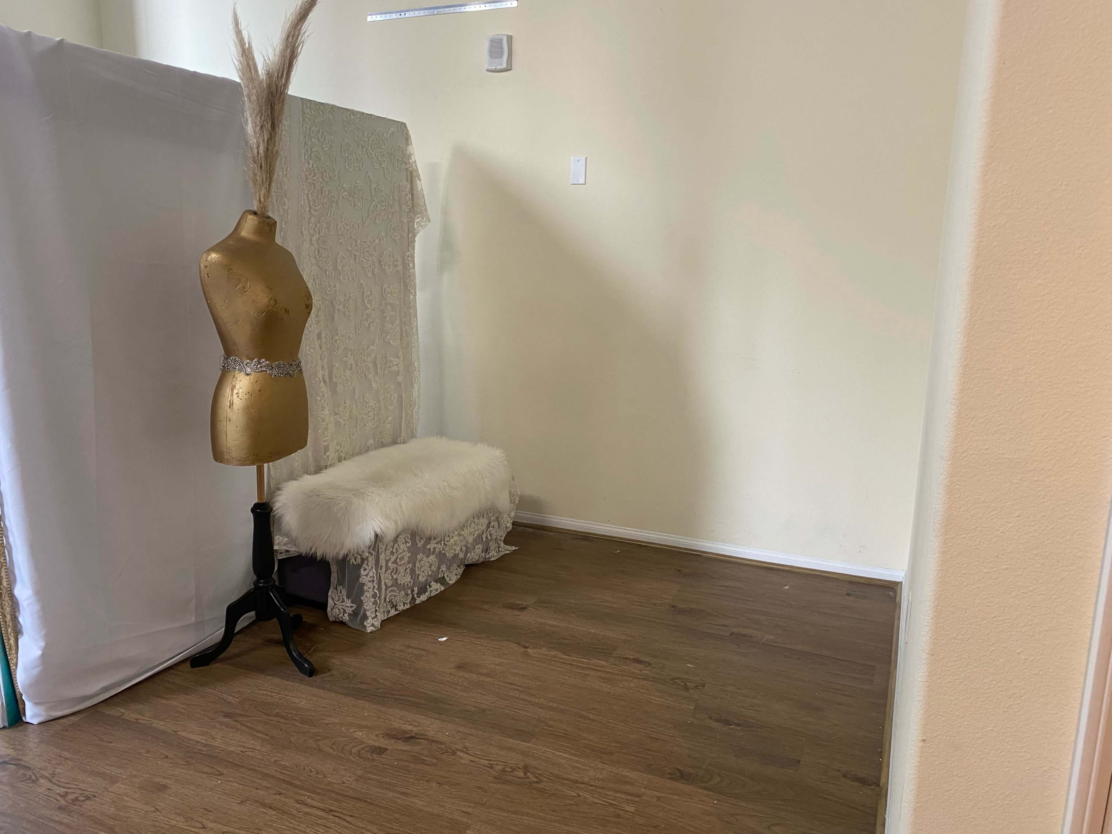 The image shows an empty room featuring a gold dress form, a white fur bench, and a backdrop of fabric.