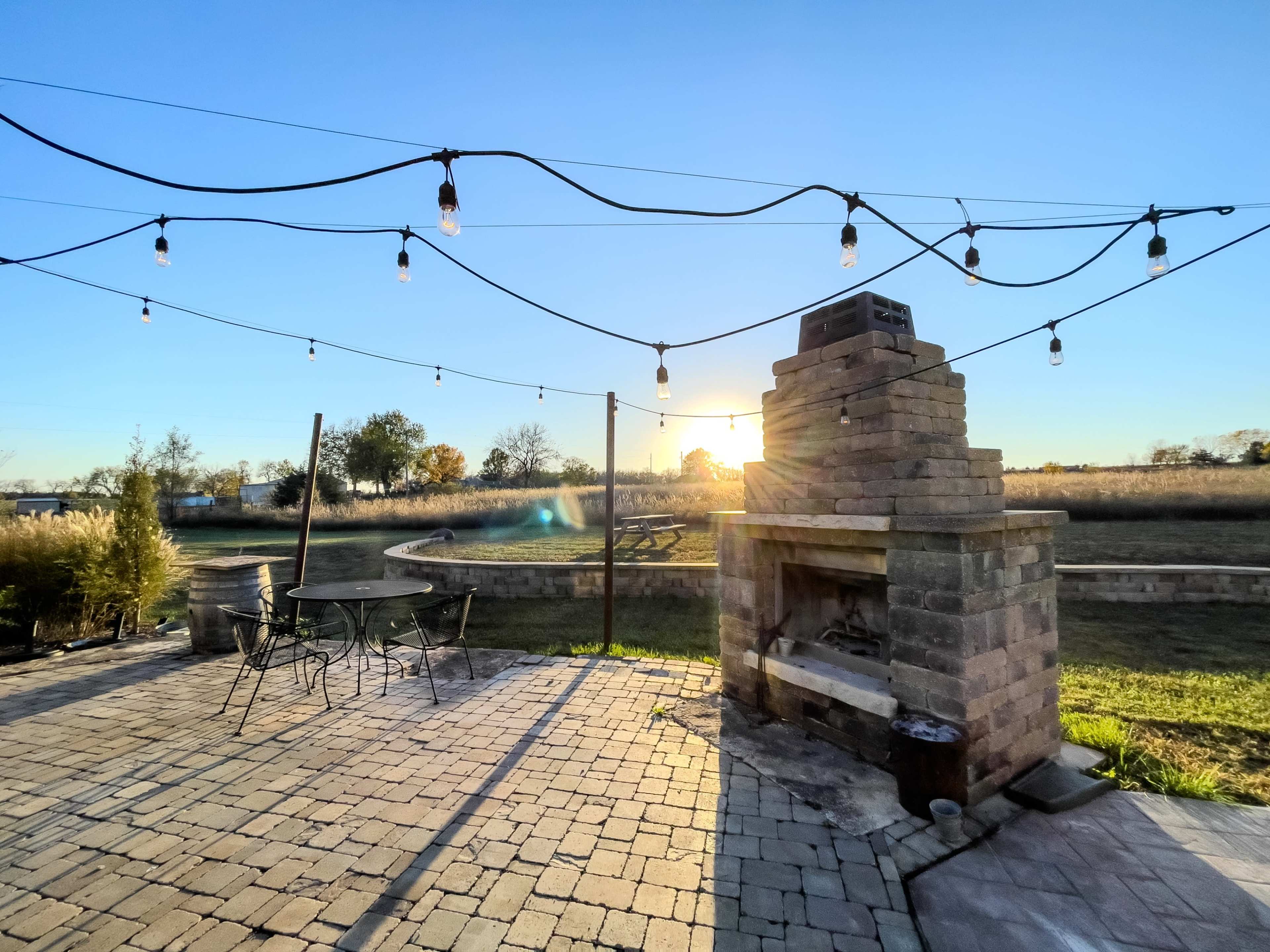A stone outdoor fireplace is illuminated by string lights as the sun sets over a grassy landscape.