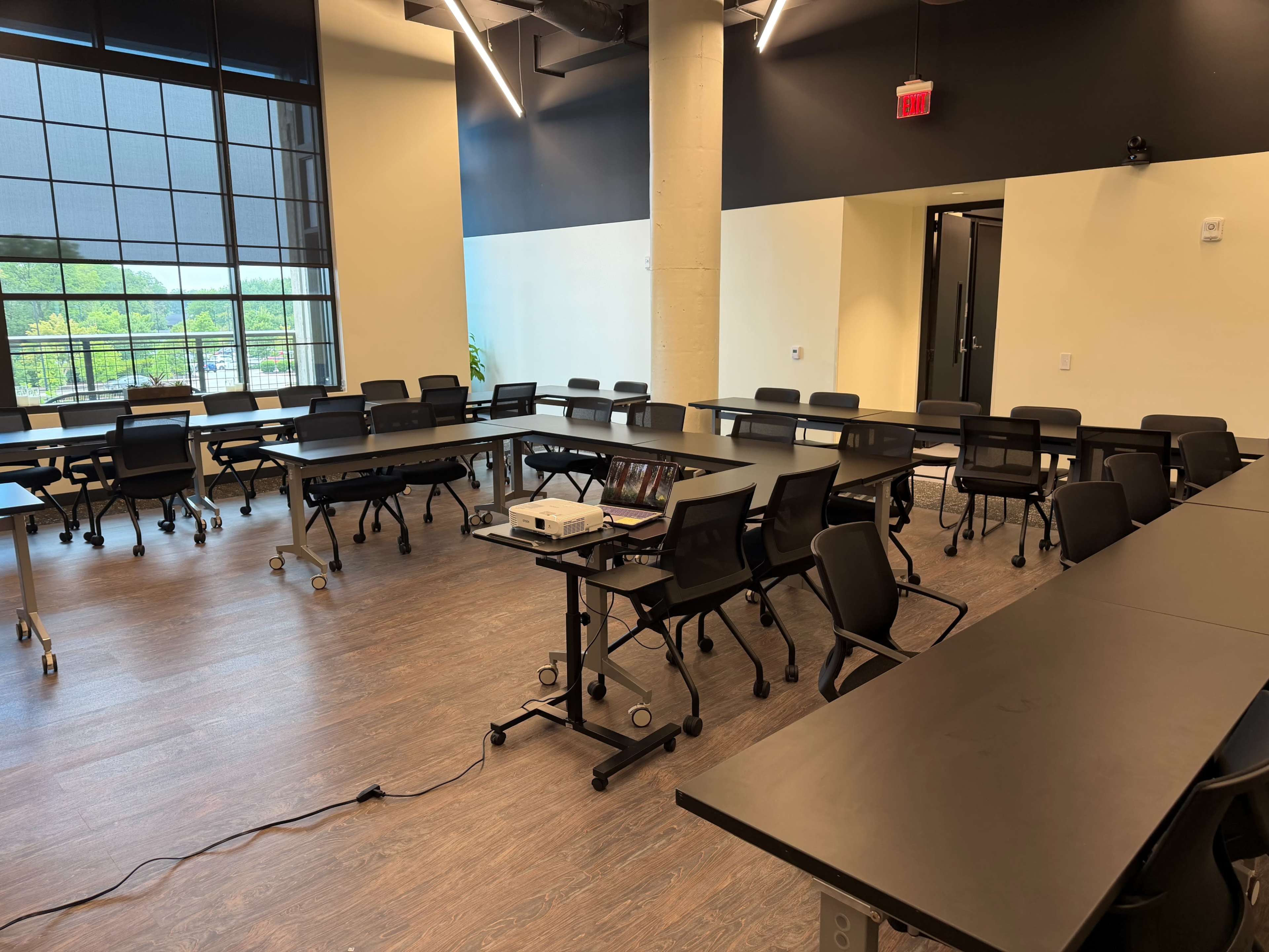 The image shows an empty classroom with multiple desks and chairs arranged in a U-shape, a projector on a table, and large windows providing natural light.