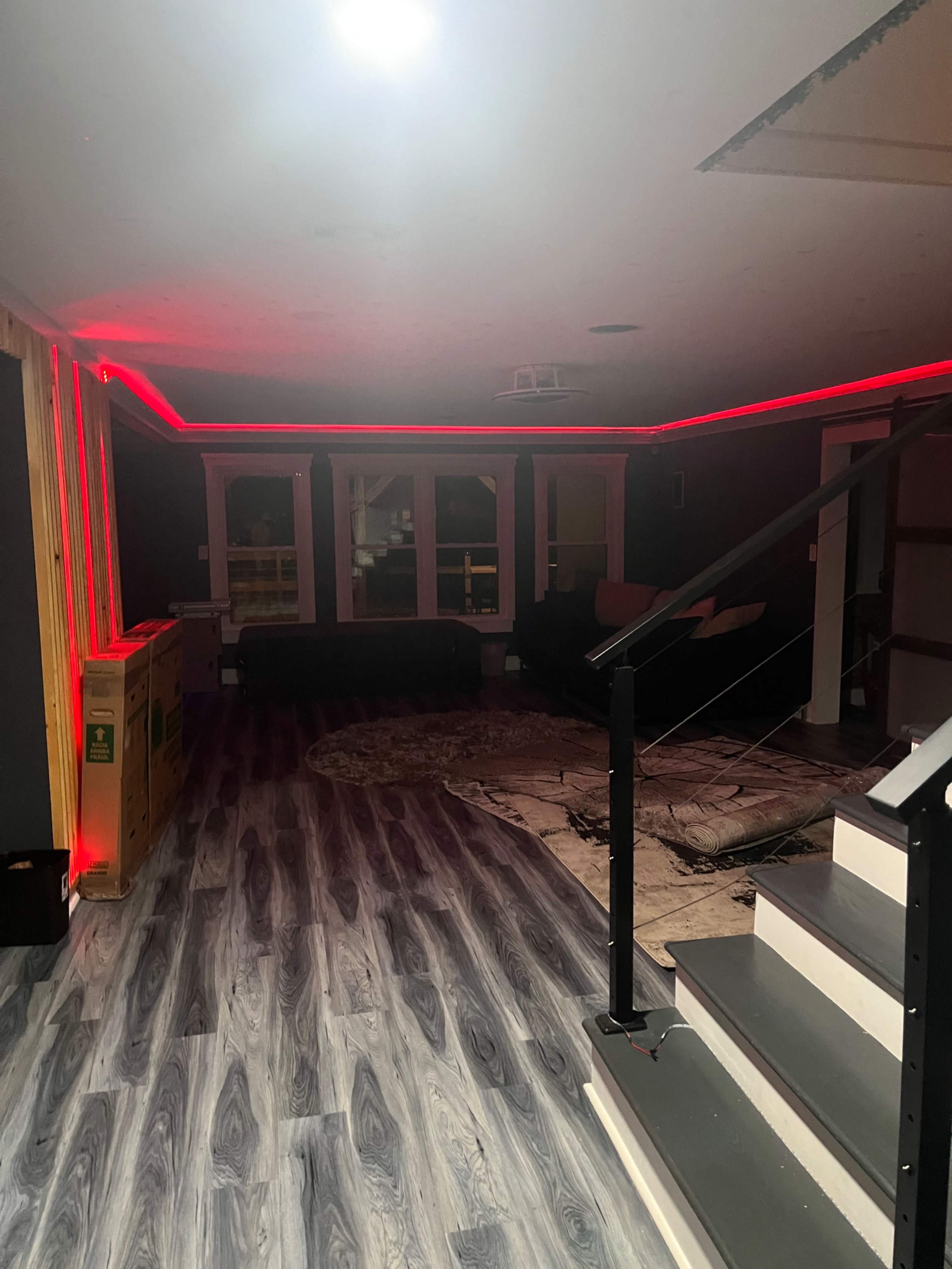 A dimly lit room with modern flooring, featuring red LED strip lighting along the walls and a staircase leading to an upper level.