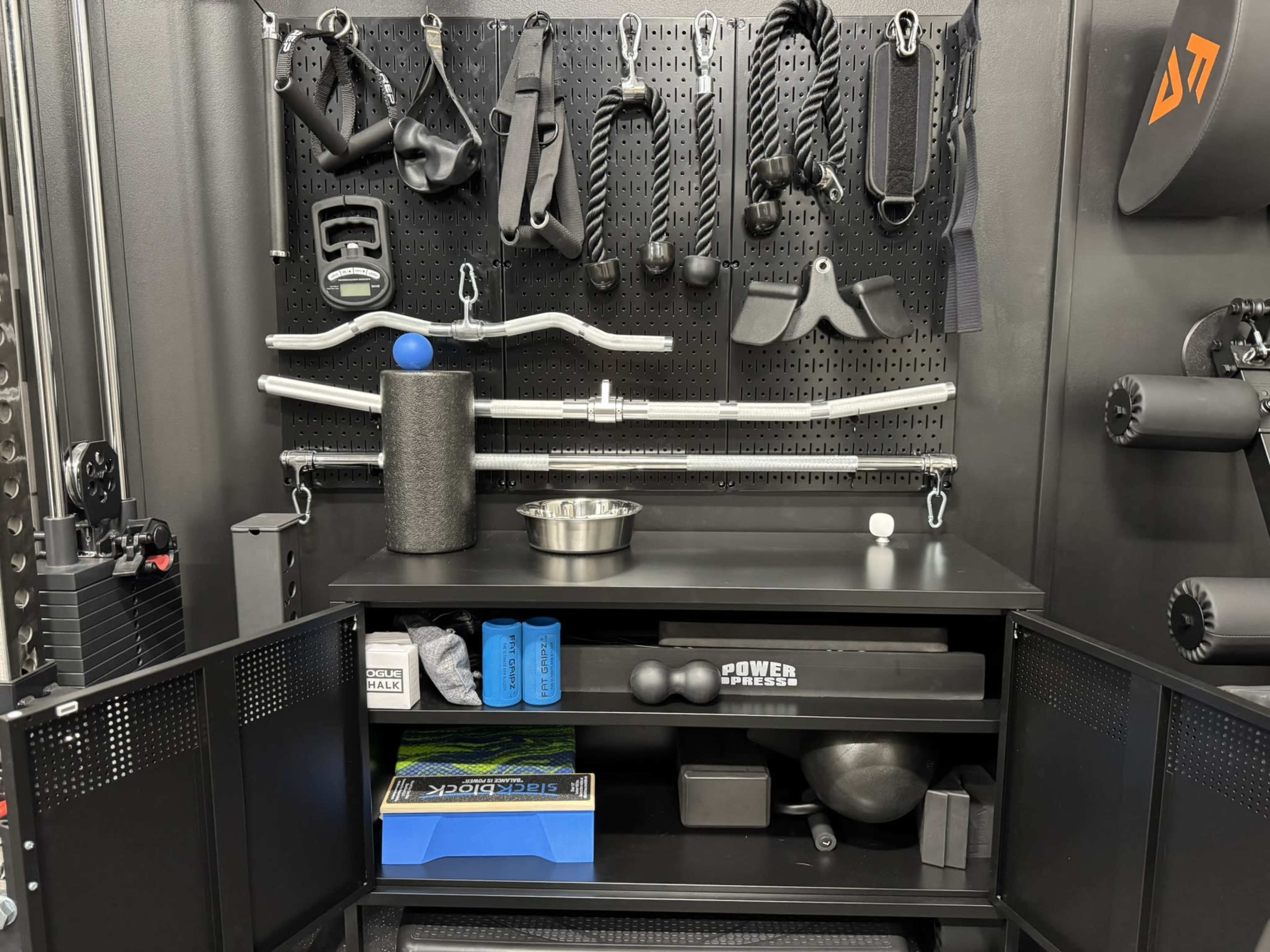 The image shows a fitness equipment storage area with various weights, resistance bands, a foam roller, and exercise accessories neatly organized on a wall-mounted pegboard and a black cabinet.