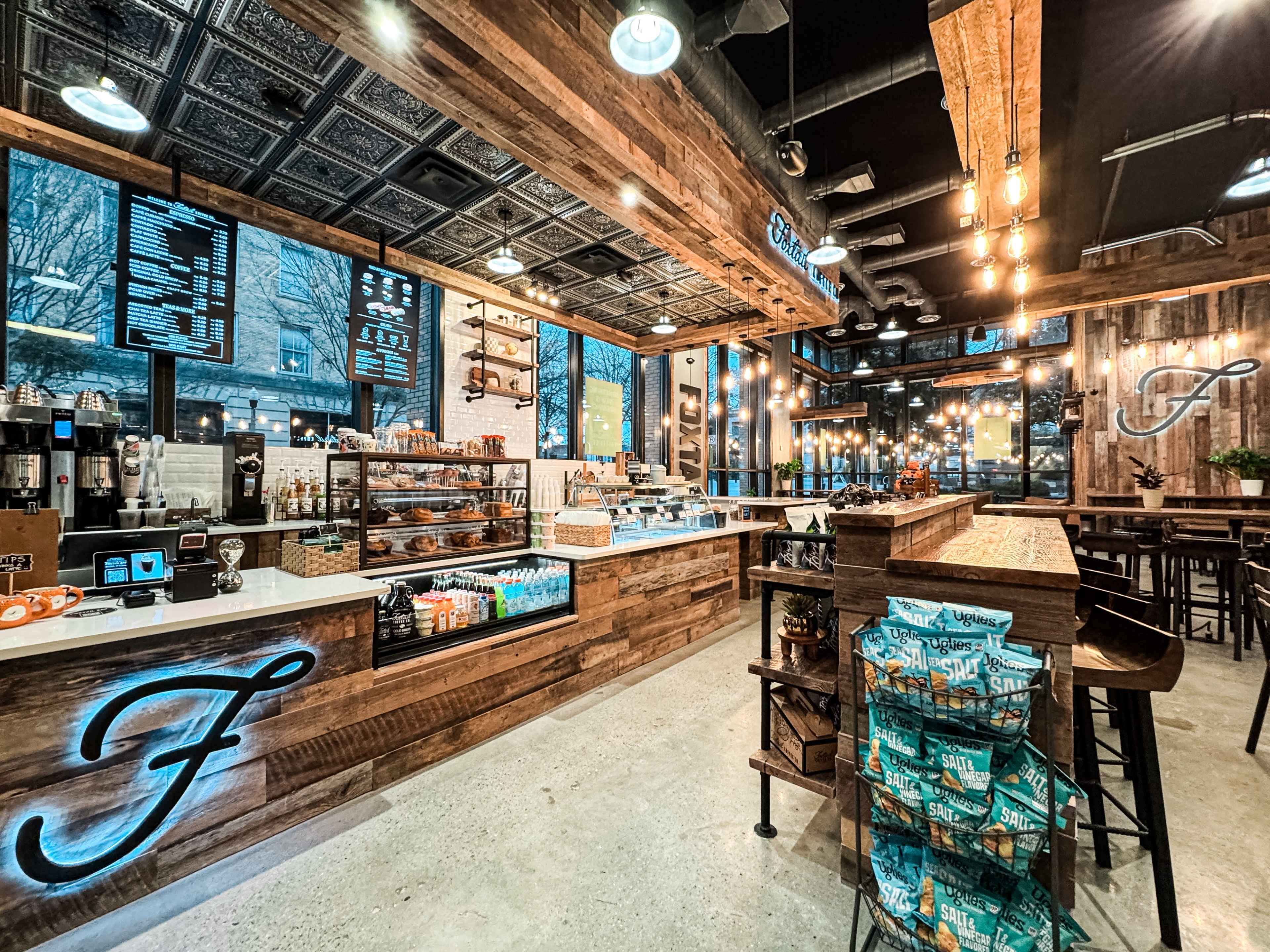 The image shows a modern cafe interior featuring wooden decor, a counter with an array of pastries and drinks, and seating arrangements with illuminated signage.