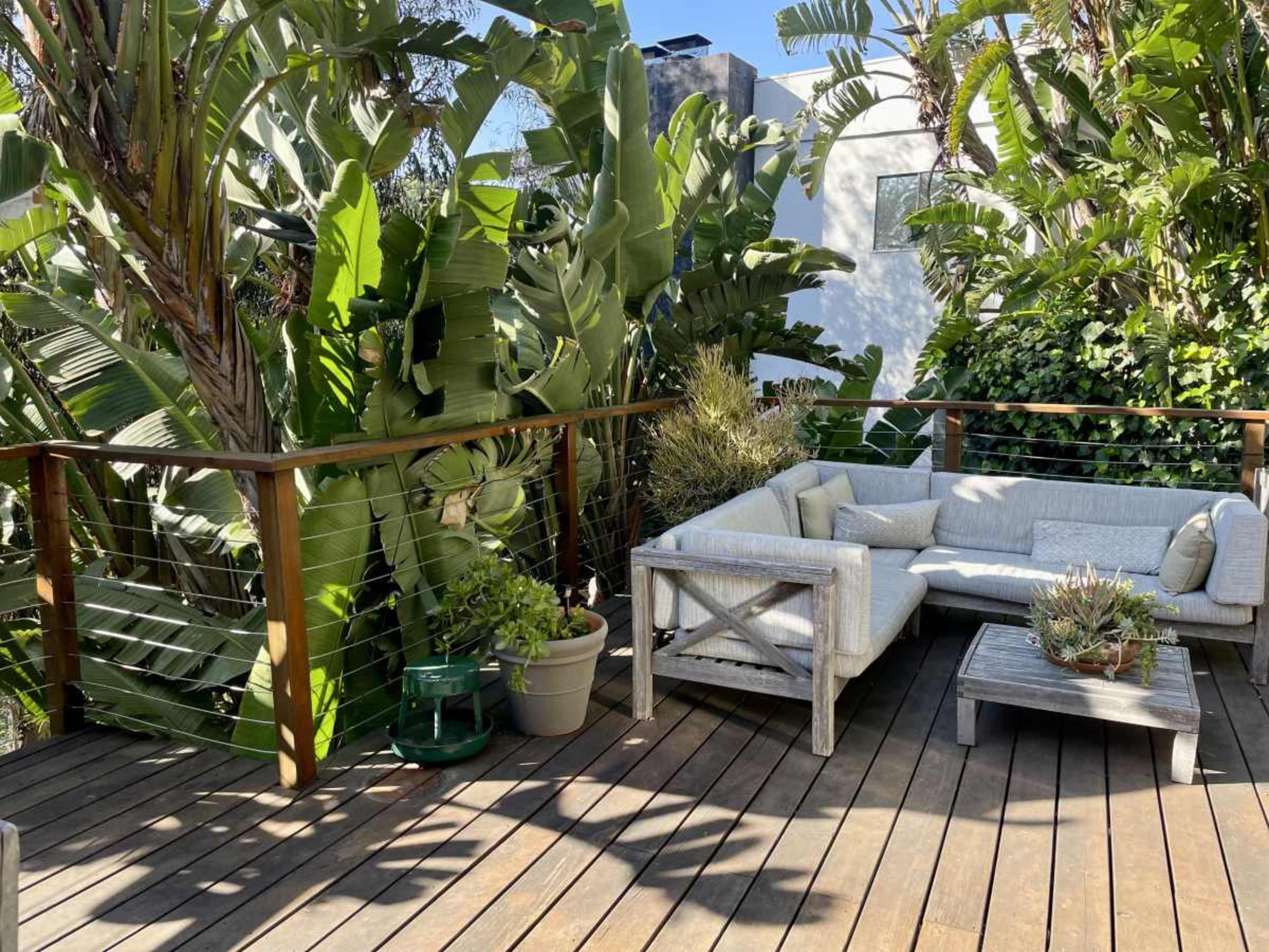 A seating area with a white sectional sofa and a small coffee table is surrounded by lush greenery on a wooden deck.