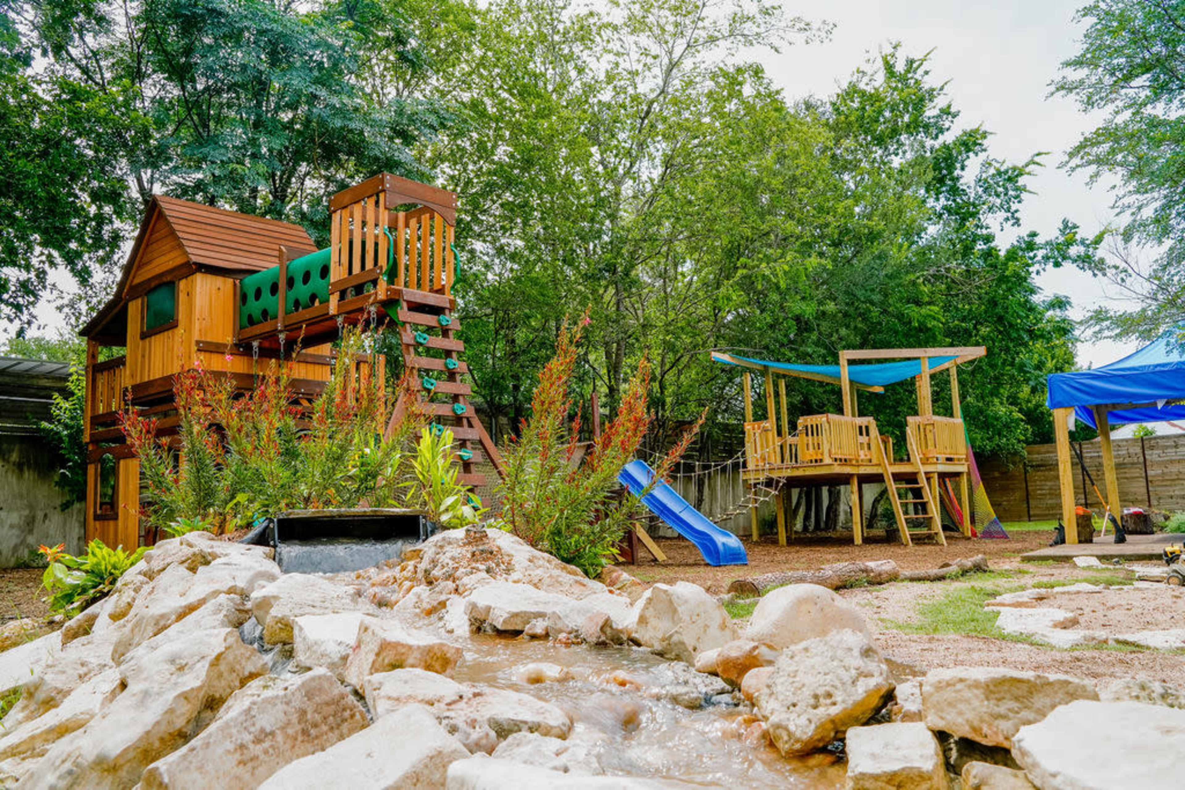 The image features a wooden playset with a slide, surrounded by greenery, alongside another wooden structure in a backyard setting.
