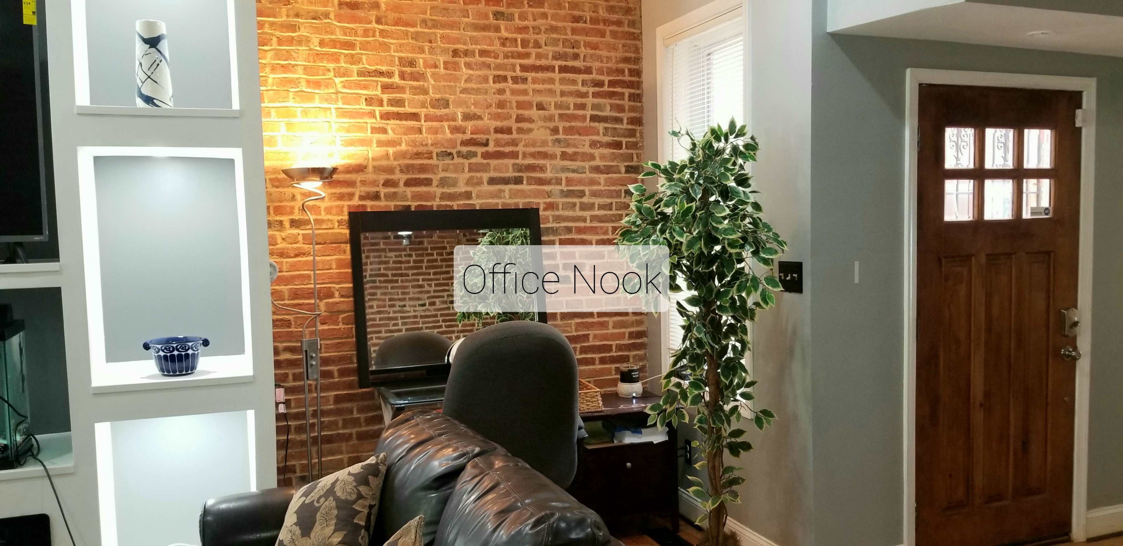 A cozy office nook with a brick wall, a desk mirror, and a chair beside a large plant.