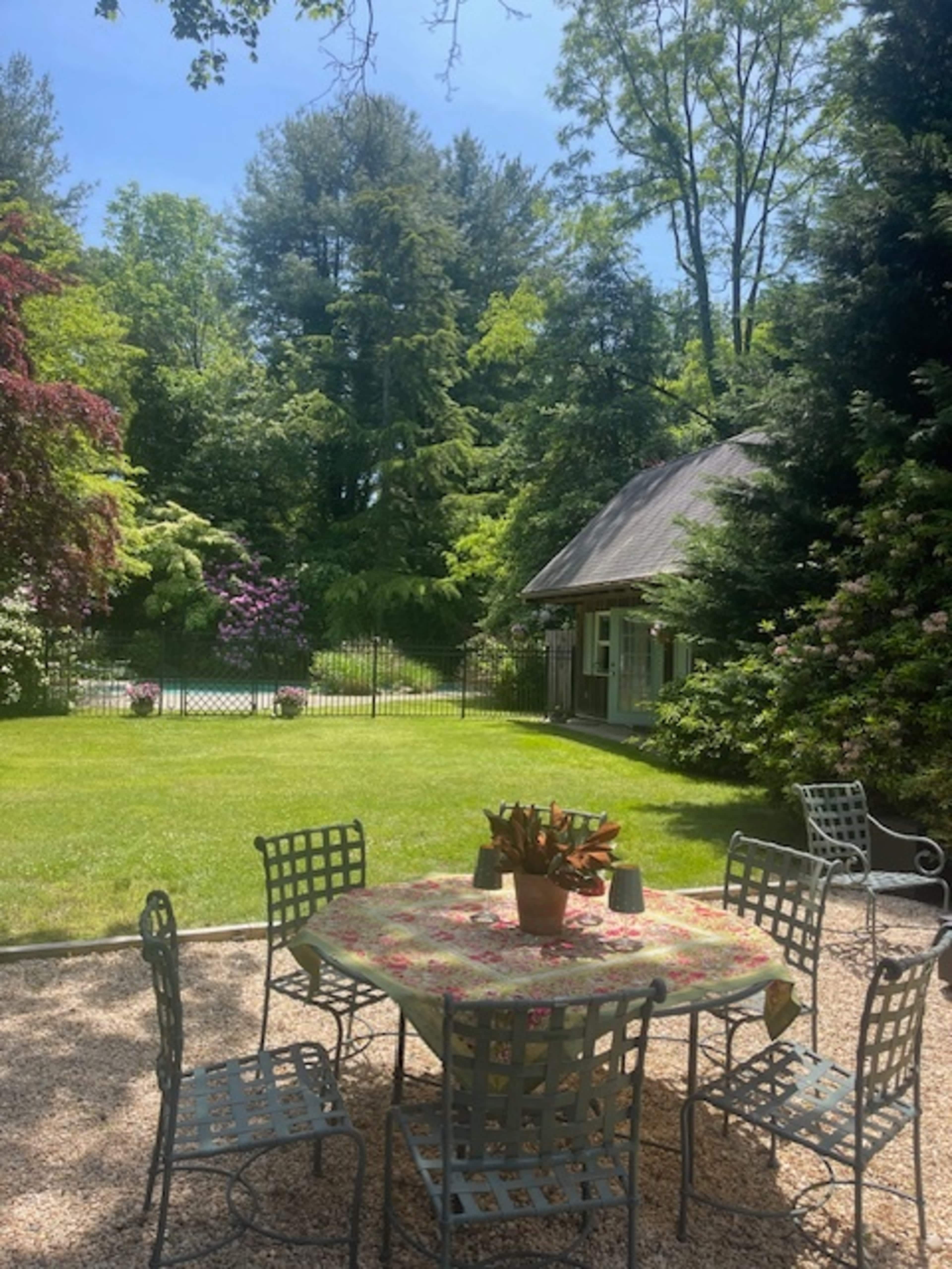 A round table surrounded by metal chairs sits on a gravel patio, with lush greenery and a small building visible in the background.
