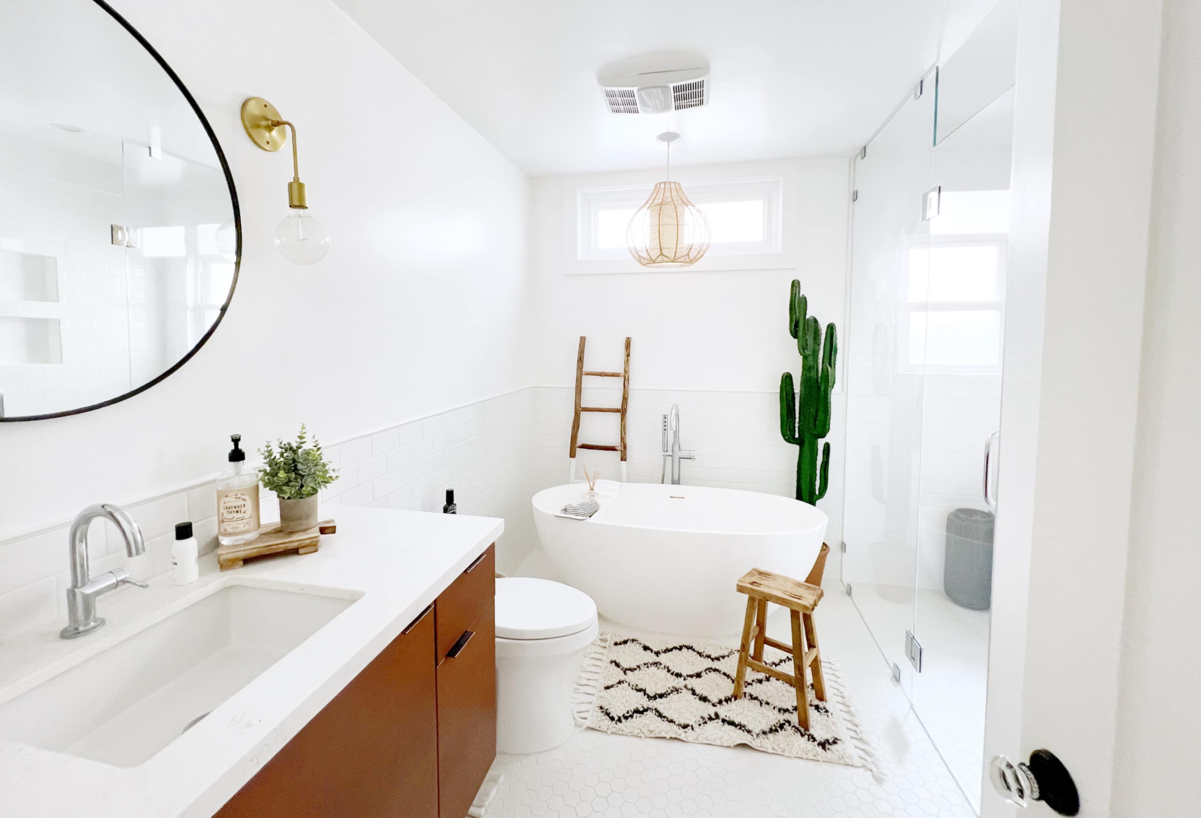The bathroom features a freestanding bathtub, a modern sink with wooden cabinetry, and a tall cactus next to a ladder, all set against a clean white backdrop.