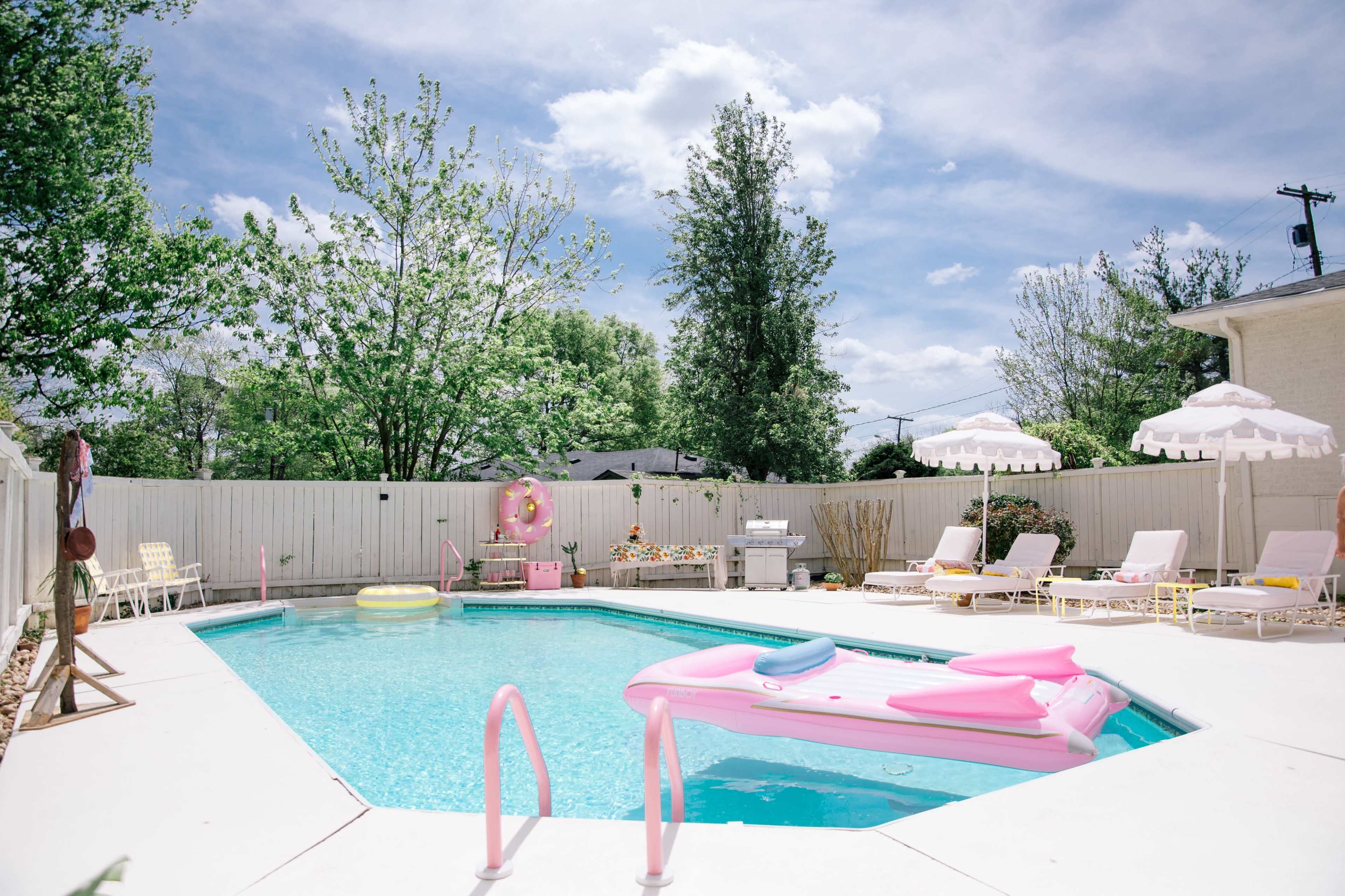 A backyard pool area features a turquoise pool with a pink float, surrounded by lounge chairs and umbrellas under a partly cloudy sky.