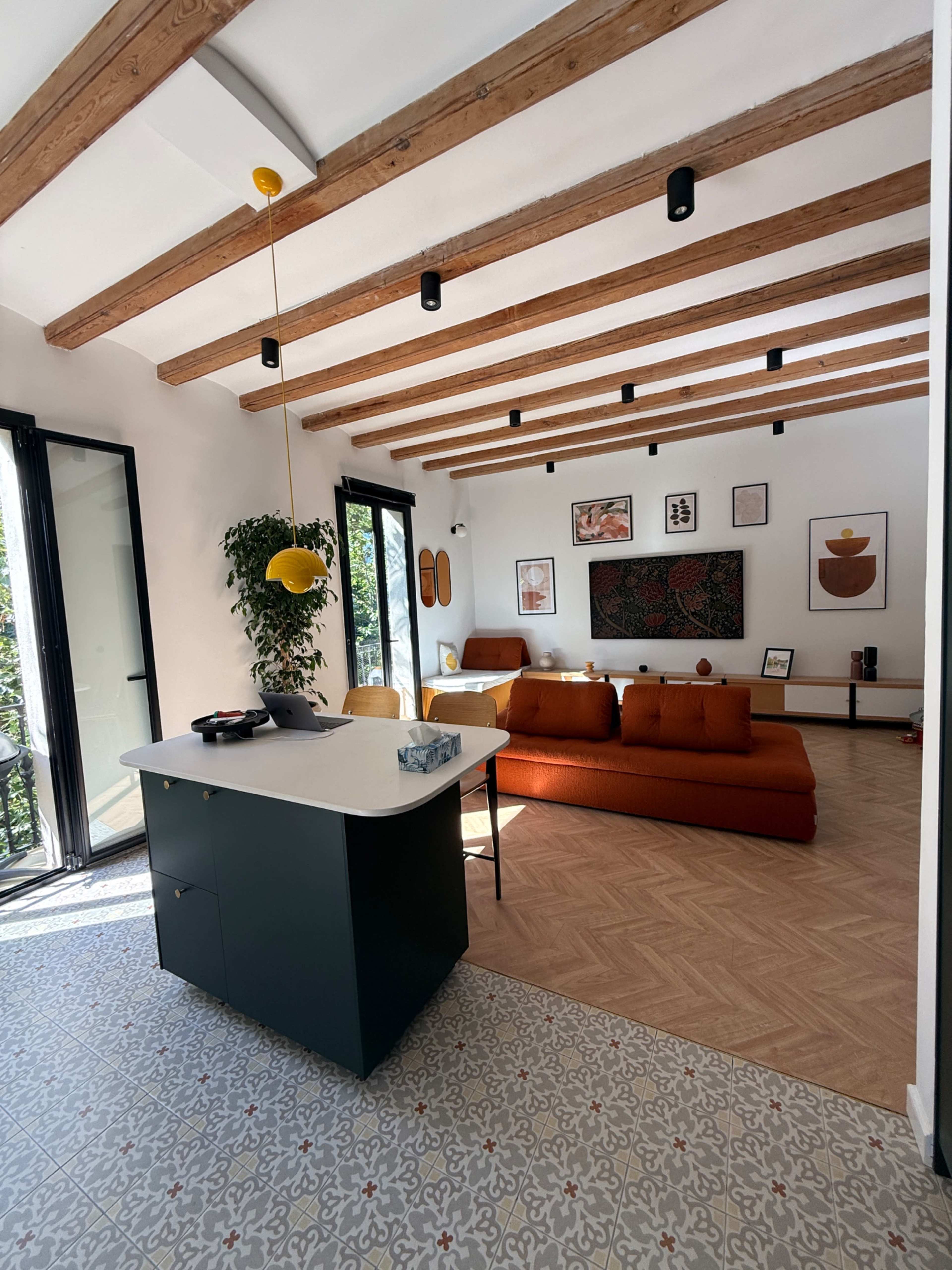 The image shows a modern living space with a kitchen island, an orange sofa, and exposed wooden beams on the ceiling.
