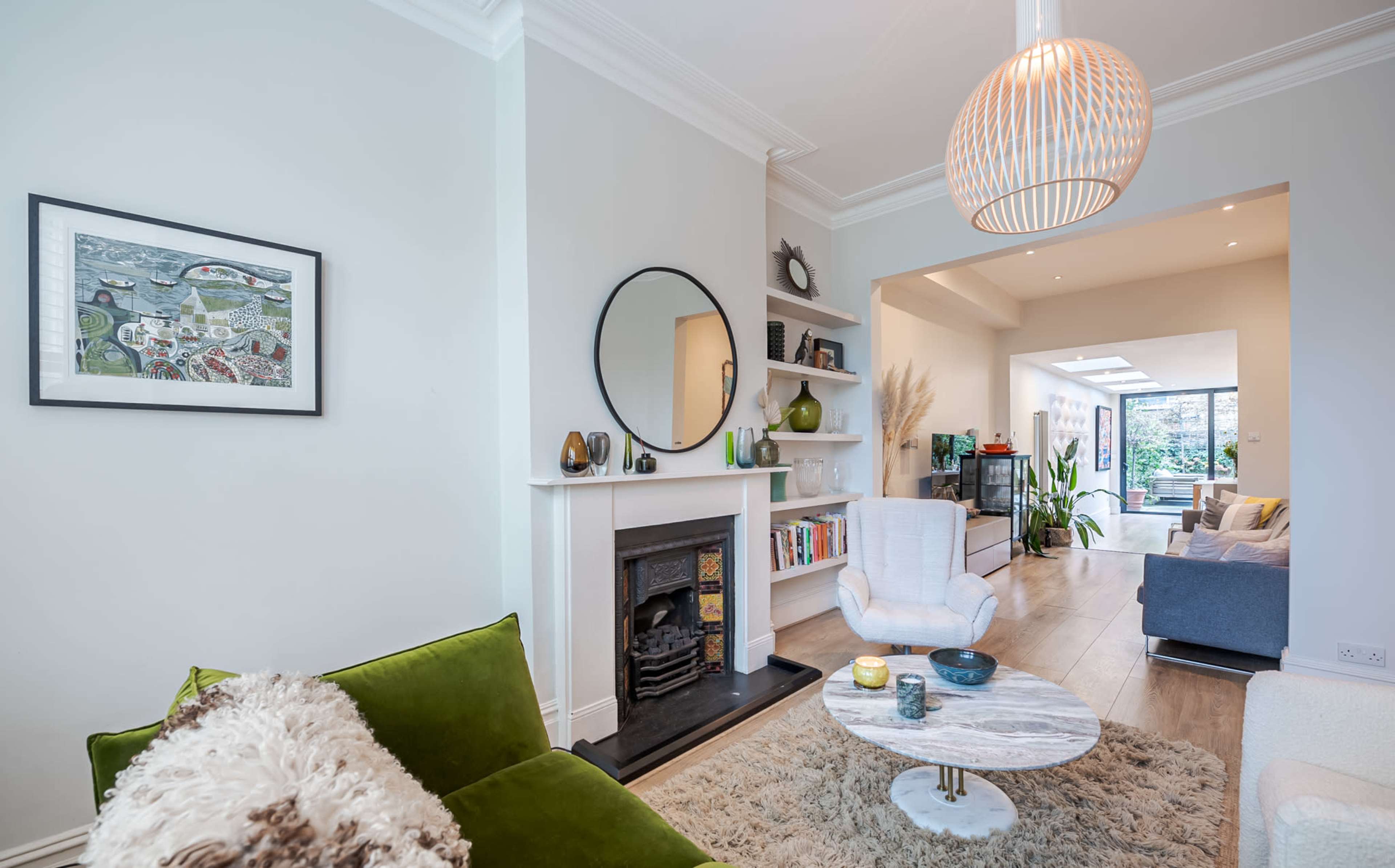 The image shows a modern living room featuring a green sofa, a round coffee table, a framed artwork on the wall, and a fireplace with a circular mirror above it.
