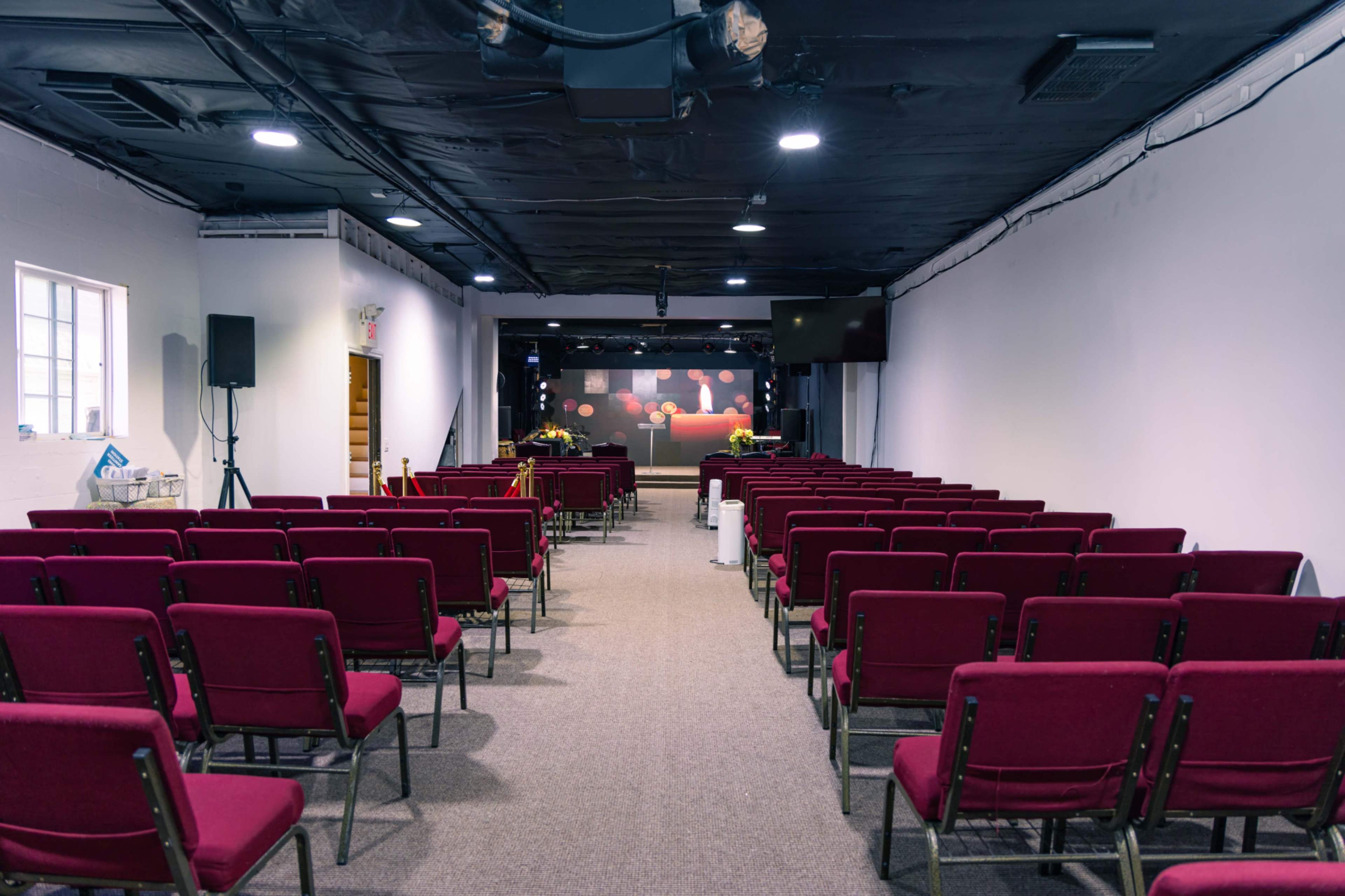 An empty hall with rows of red chairs facing a stage at the front, illuminated by overhead lights.