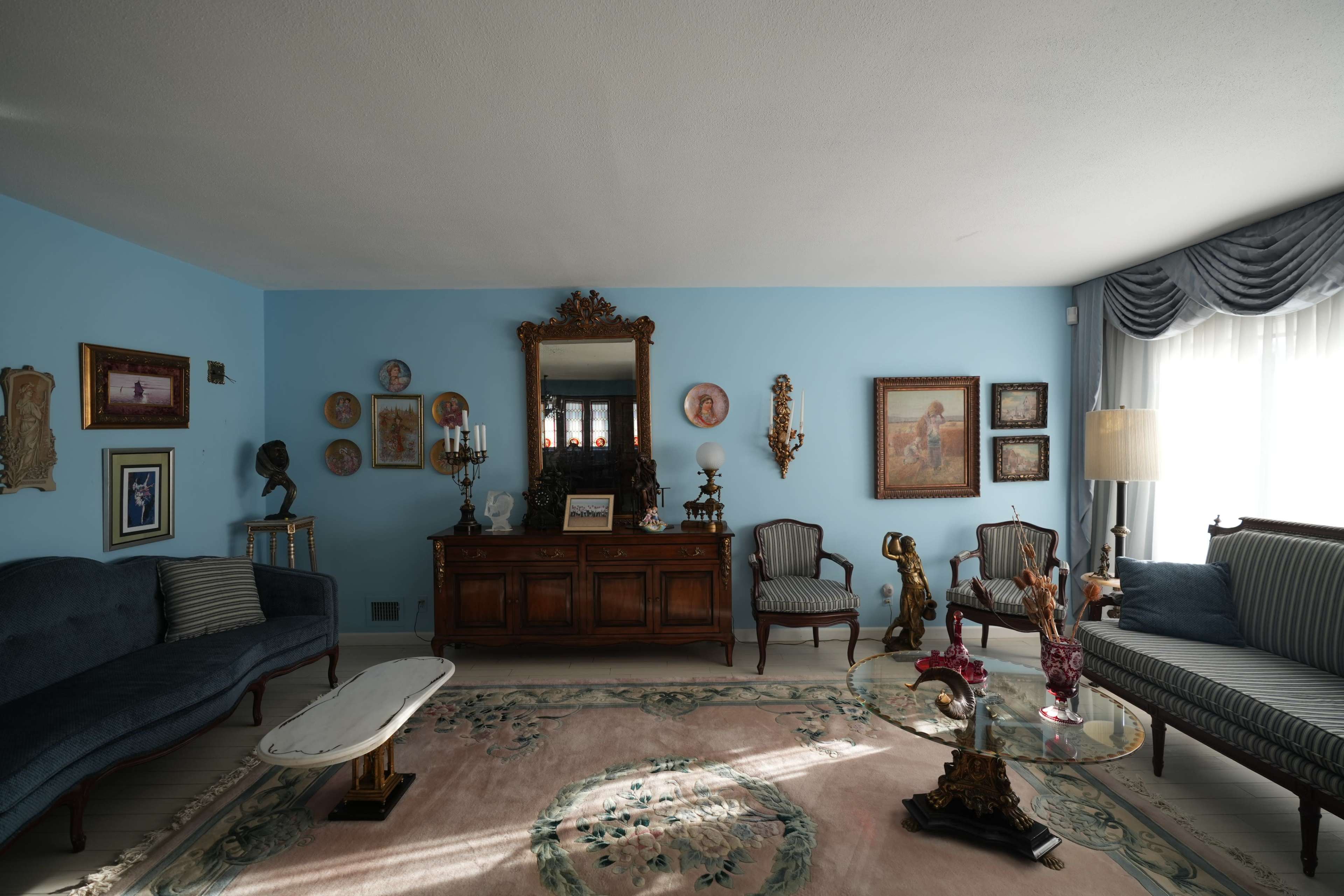 The room features a blue wall, a variety of framed artworks, a wooden sideboard, and vintage furniture arranged around a patterned rug.