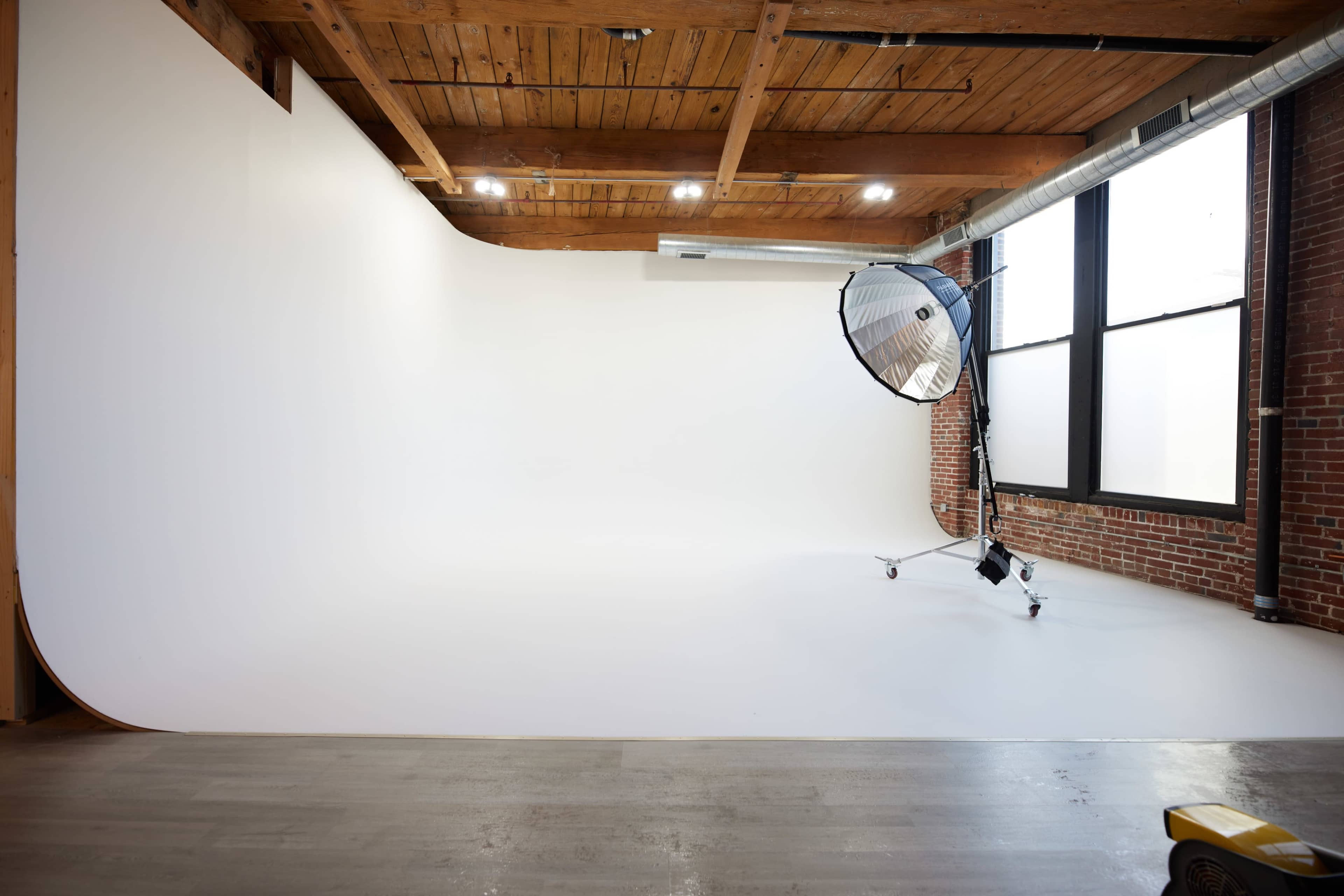 The image shows a photography studio with a white backdrop, a large softbox light on wheels, and exposed wooden beams overhead.